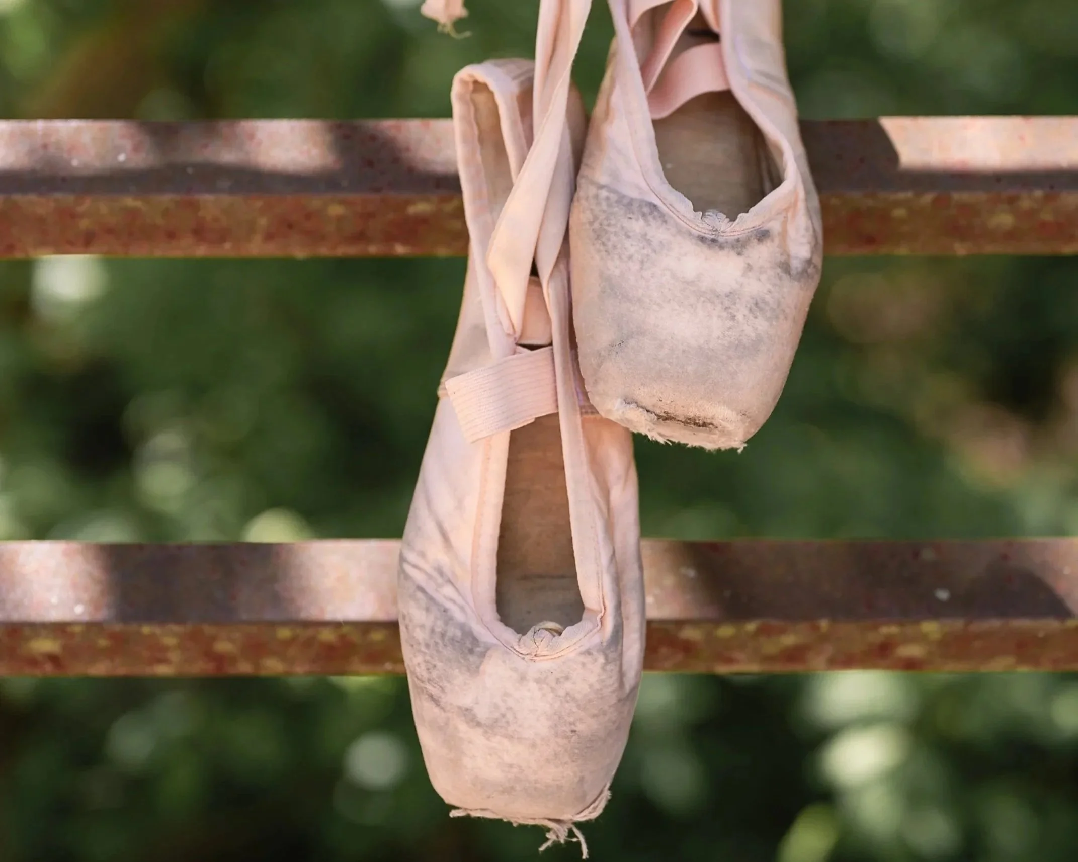 A pair of worn pink ballet pointe shoes hanging from a rusty metal railing outdoors.
