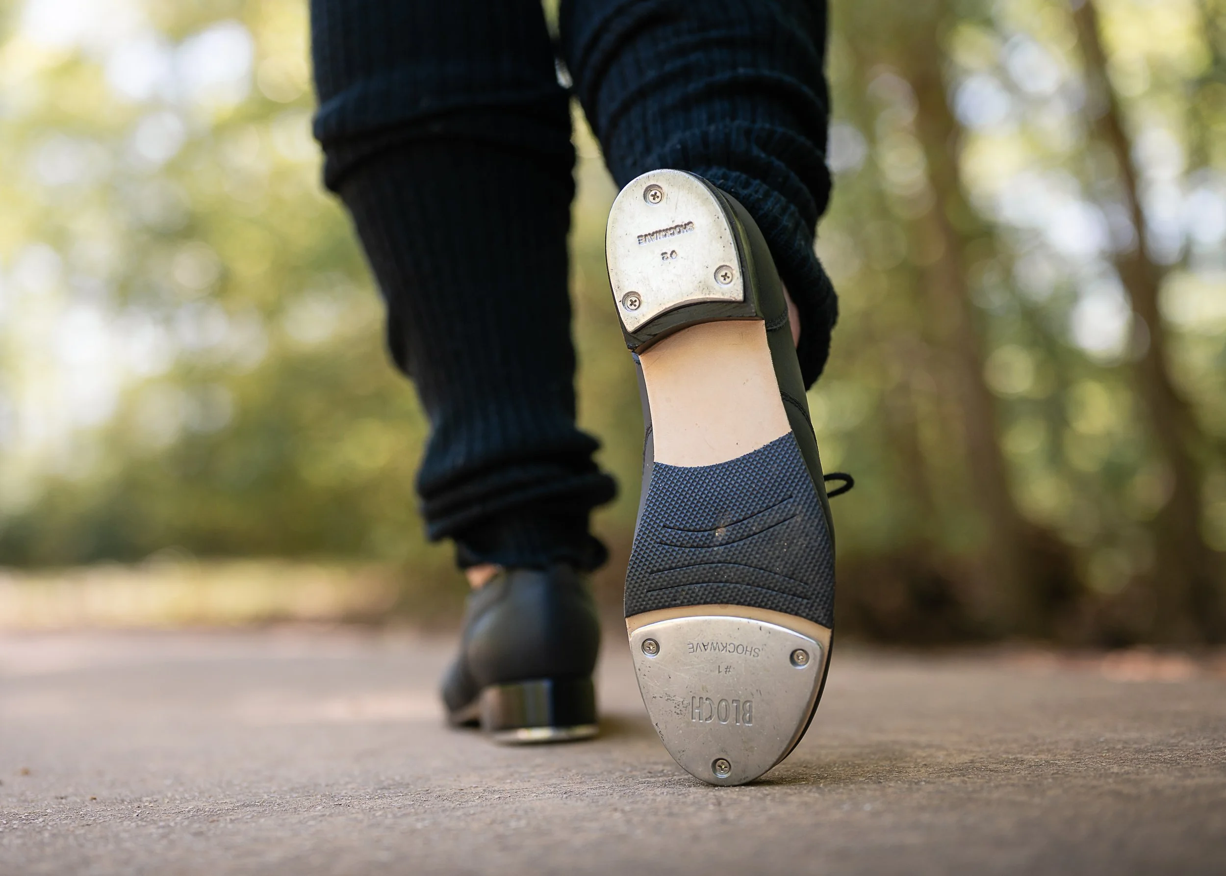 A person wearing dark pants and tap shoes taking a step away, with a blurred background of trees.