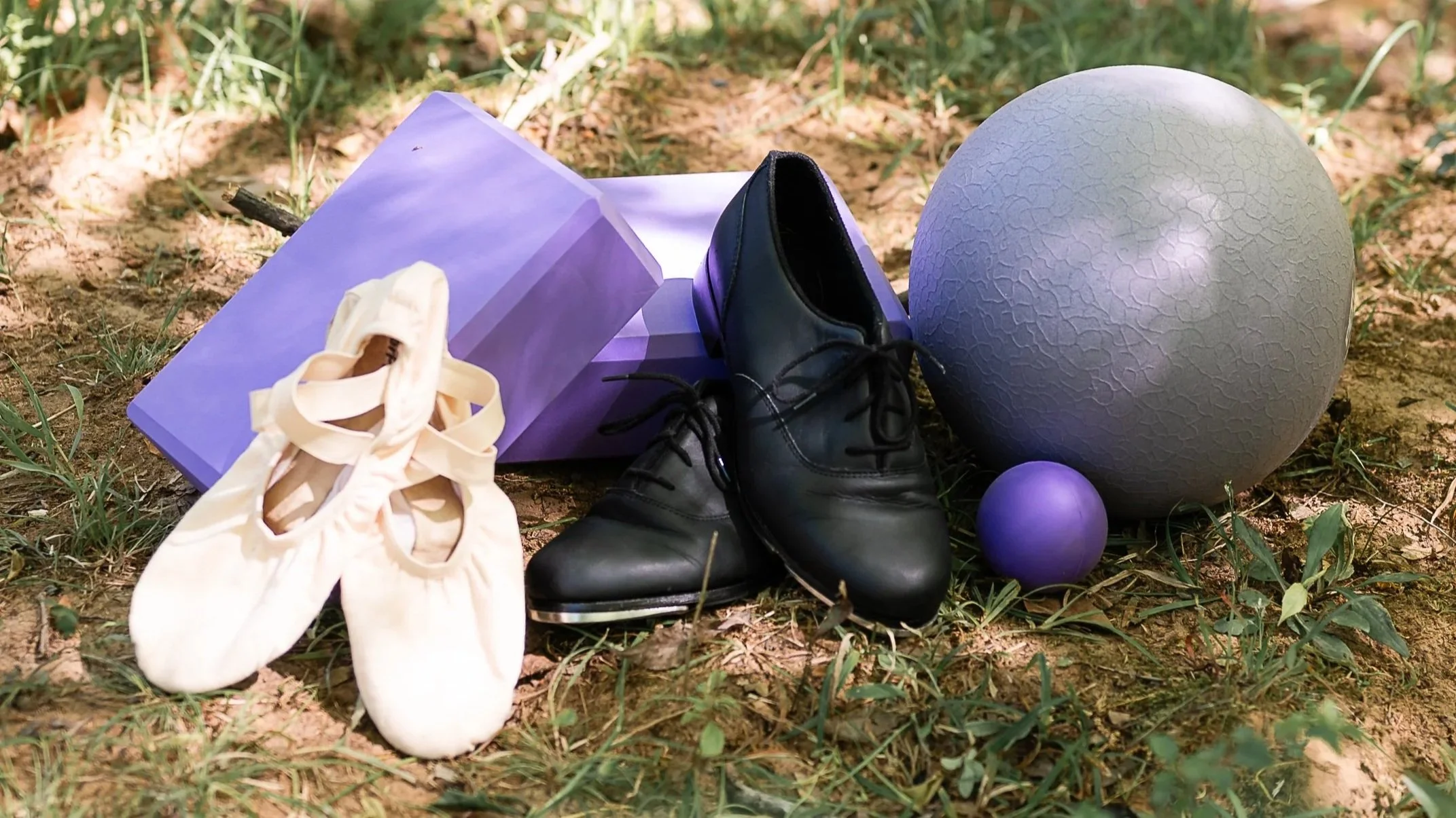 A collection of dance or ballet-related items on the ground outdoors, including a pair of ballet shoes, black leather lace-up shoes, a purple yoga ball, a purple ball, a large textured spherical object, a purple mat, and a small purple object, surrounded by grass and dirt.