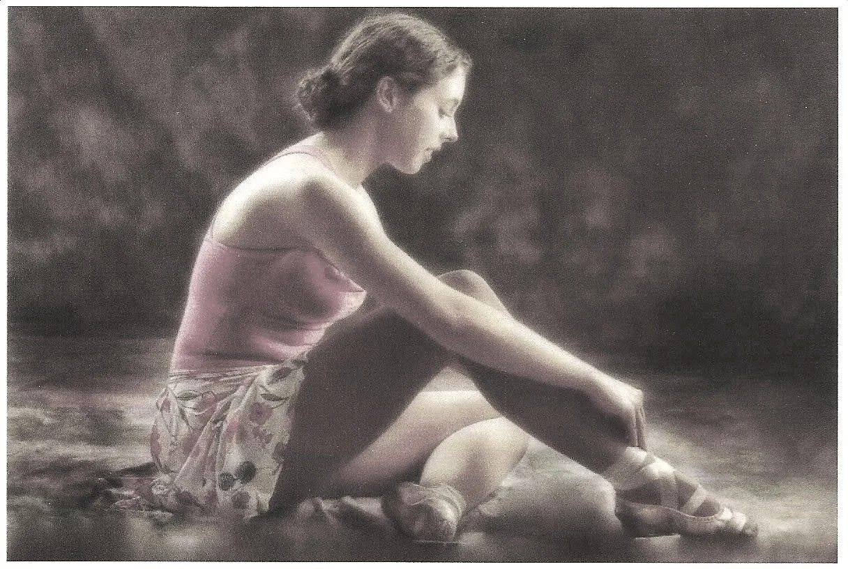 A young woman sitting in a dance studio with her legs crossed, looking down, wearing a pink leotard, floral skirt, and pointe shoes.