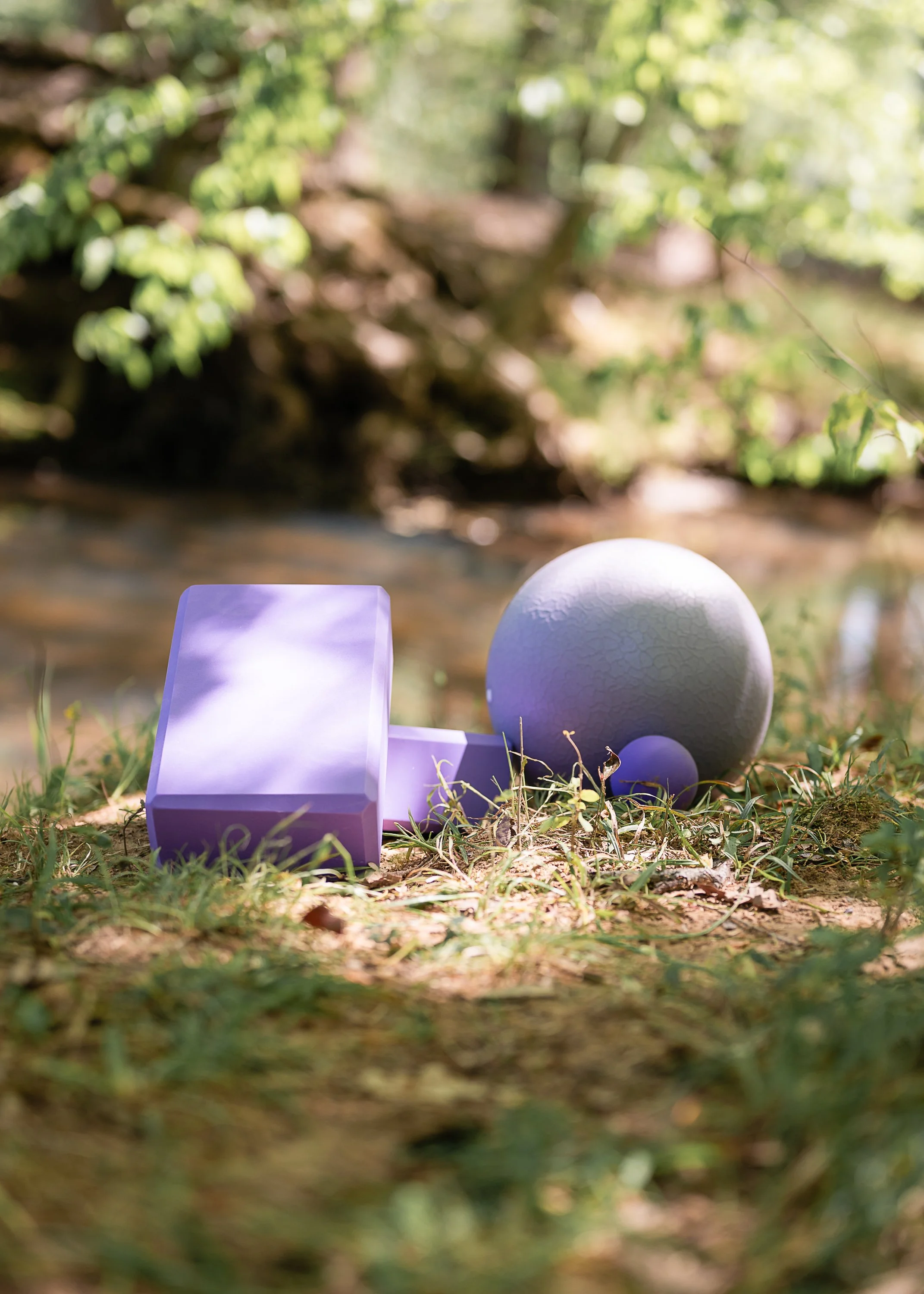 Close-up of three painted objects with purple, white, and lavender colors on grass, with trees and greenery in the blurry background.