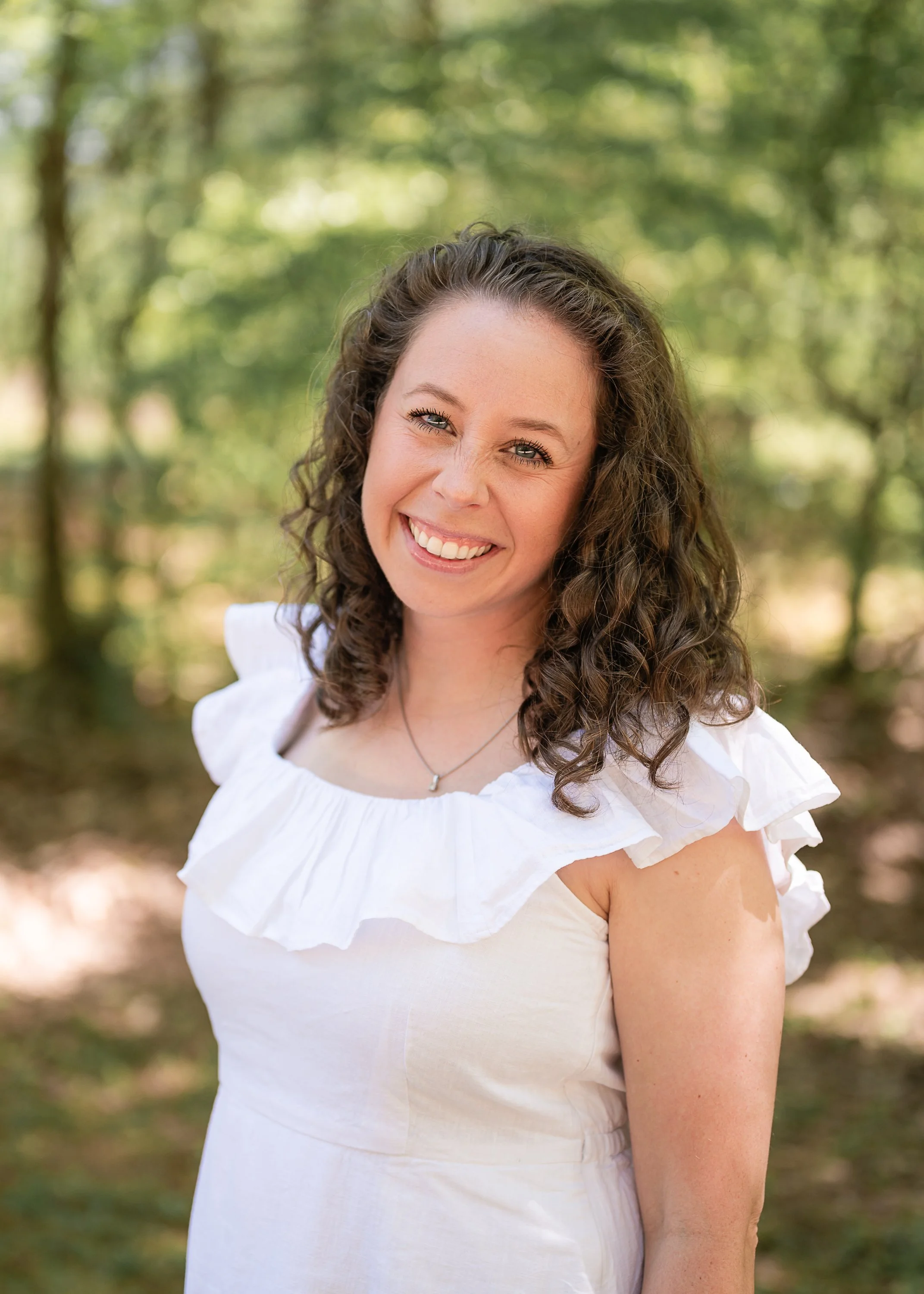 A woman with curly brown hair smiling outdoors in a sunny wooded area, wearing a white ruffled top and a necklace.