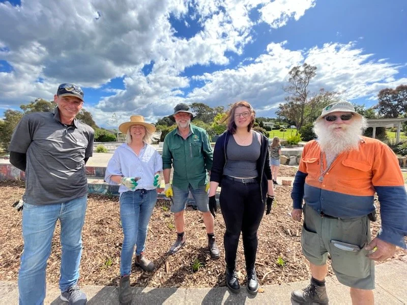 Group of five people outdoors planting trees or plants in a garden with a partly cloudy sky in the background.
