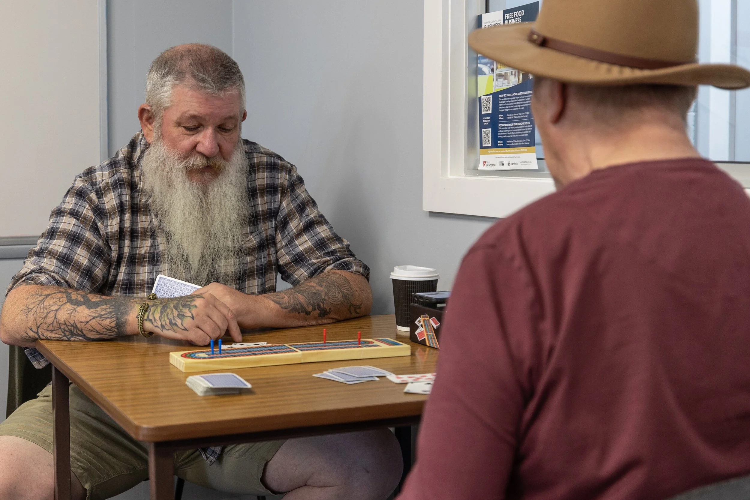 Two men playing a game of Scrabble at a table in an indoor setting. One man has a long white beard, tattoos on his arms, and is wearing a plaid shirt. The other man is wearing a wide-brimmed hat and a red shirt. There are a coffee cup, notebook, and 
