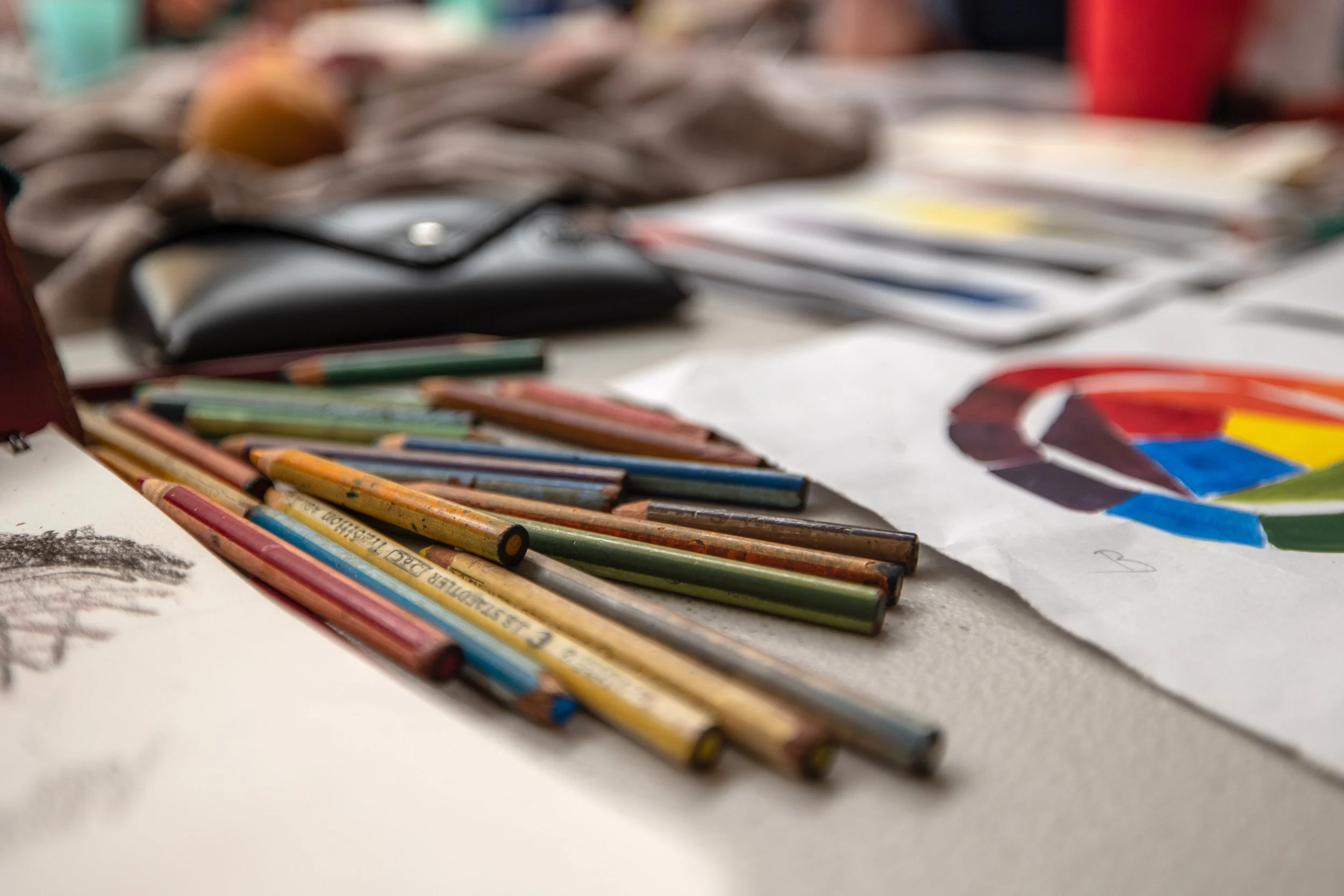Colored pencils scattered on a table with some drawings and a sketchbook in the background.