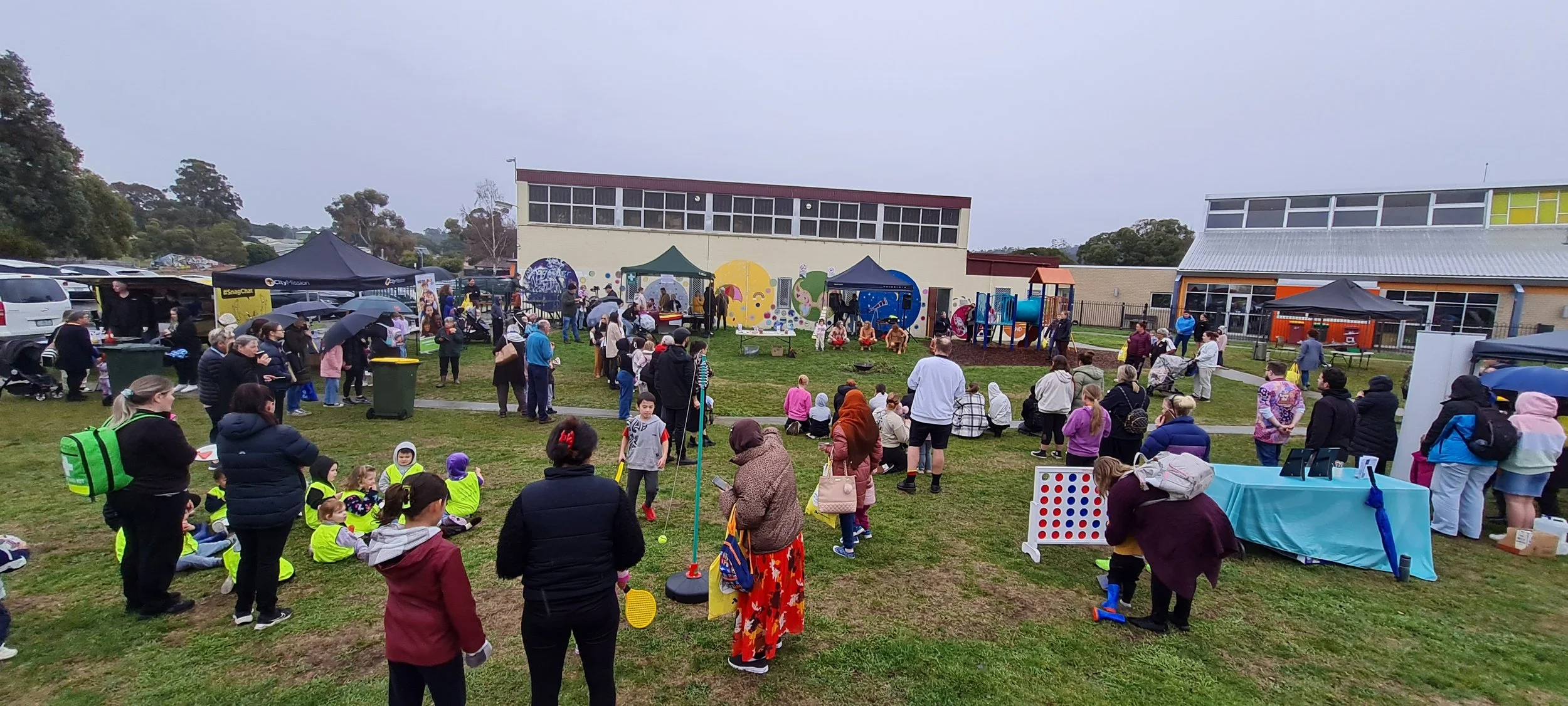 A community outdoor event at a school playground on a cloudy day, featuring people of all ages, tents, booths, a play area, and a mural wall in the background.