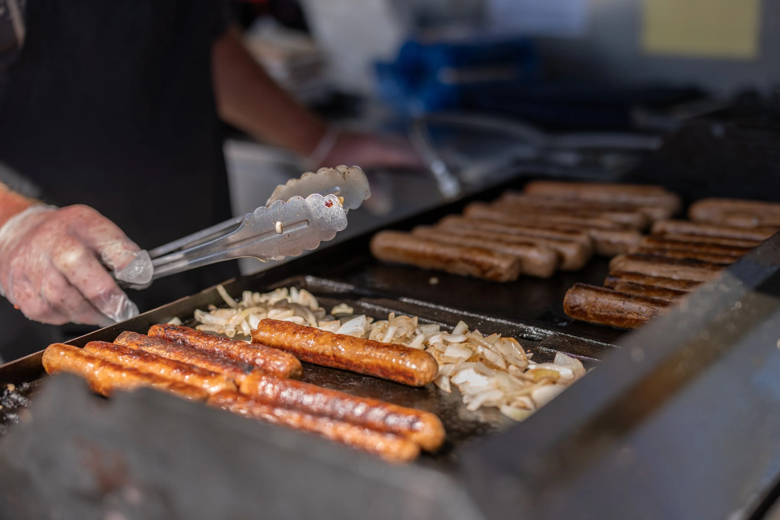 Hot dogs and onions cooking on a grill with a person using tongs to turn them.