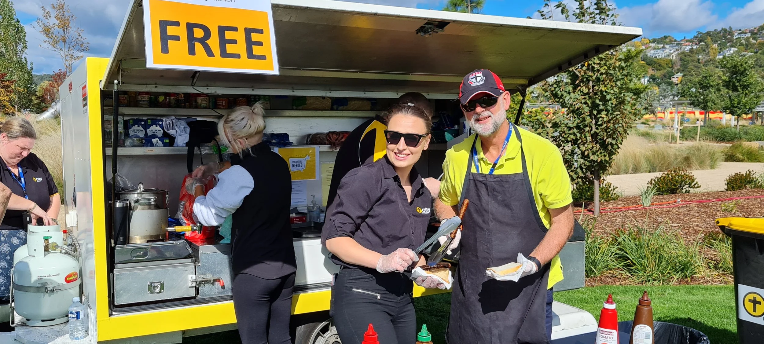 Two people smiling and serving hot dogs at a food truck with a 'FREE' sign on top, outdoors on a sunny day, with other staff members in the background and scenic greenery.