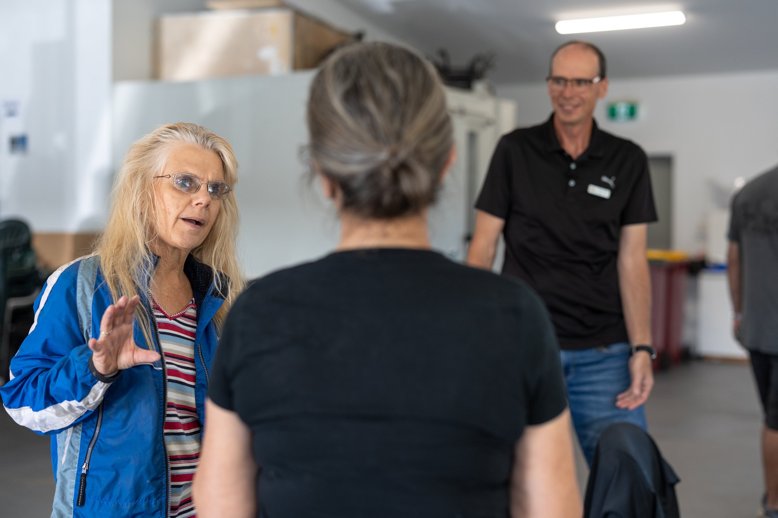 Three people engaged in a conversation indoors, with a woman speaking and gesturing, and two others listening, in a casual setting.