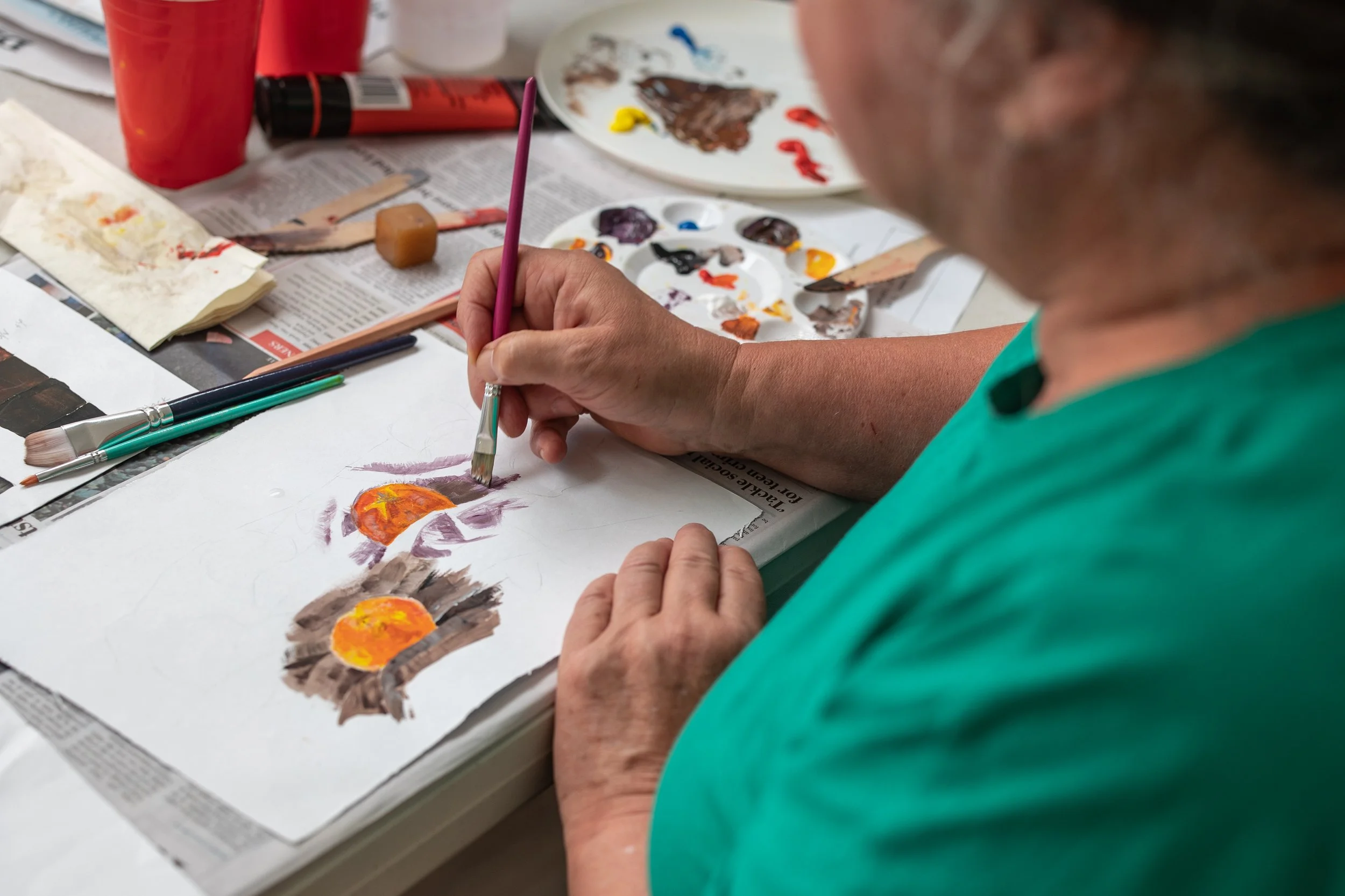 Person painting a picture of three orange and yellow flowers on white paper with watercolor paints, surrounded by brushes, paint palettes, and art supplies on a newspaper-covered table.
