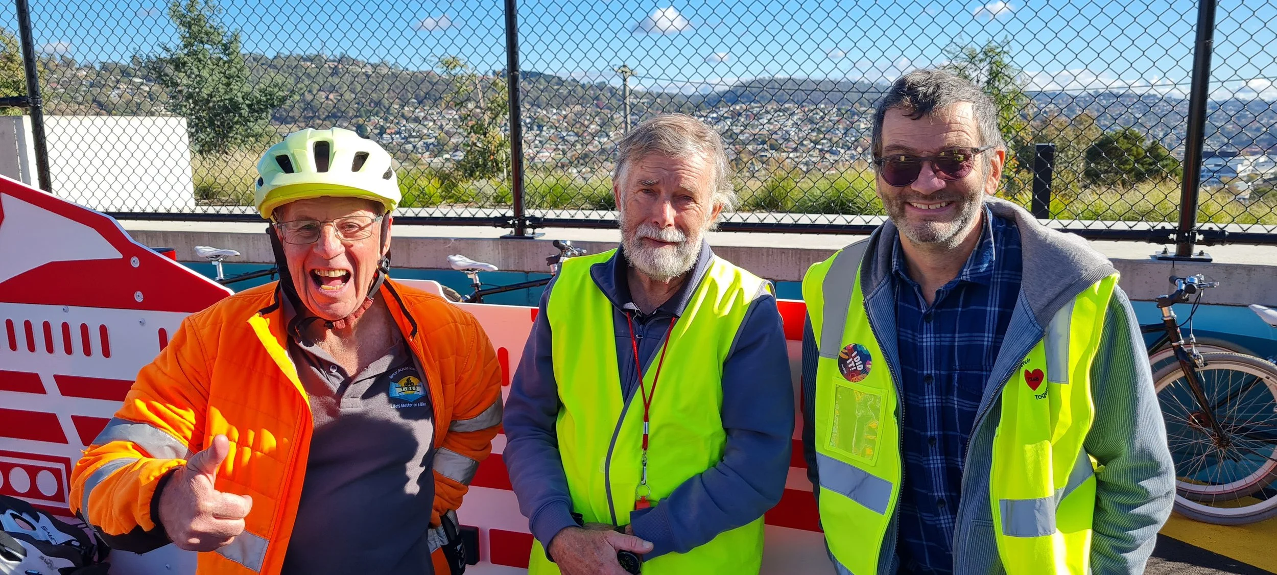 Three men smiling and standing together outdoors, wearing cycling gear and high-visibility vests, with bicycles and a cityscape in the background.