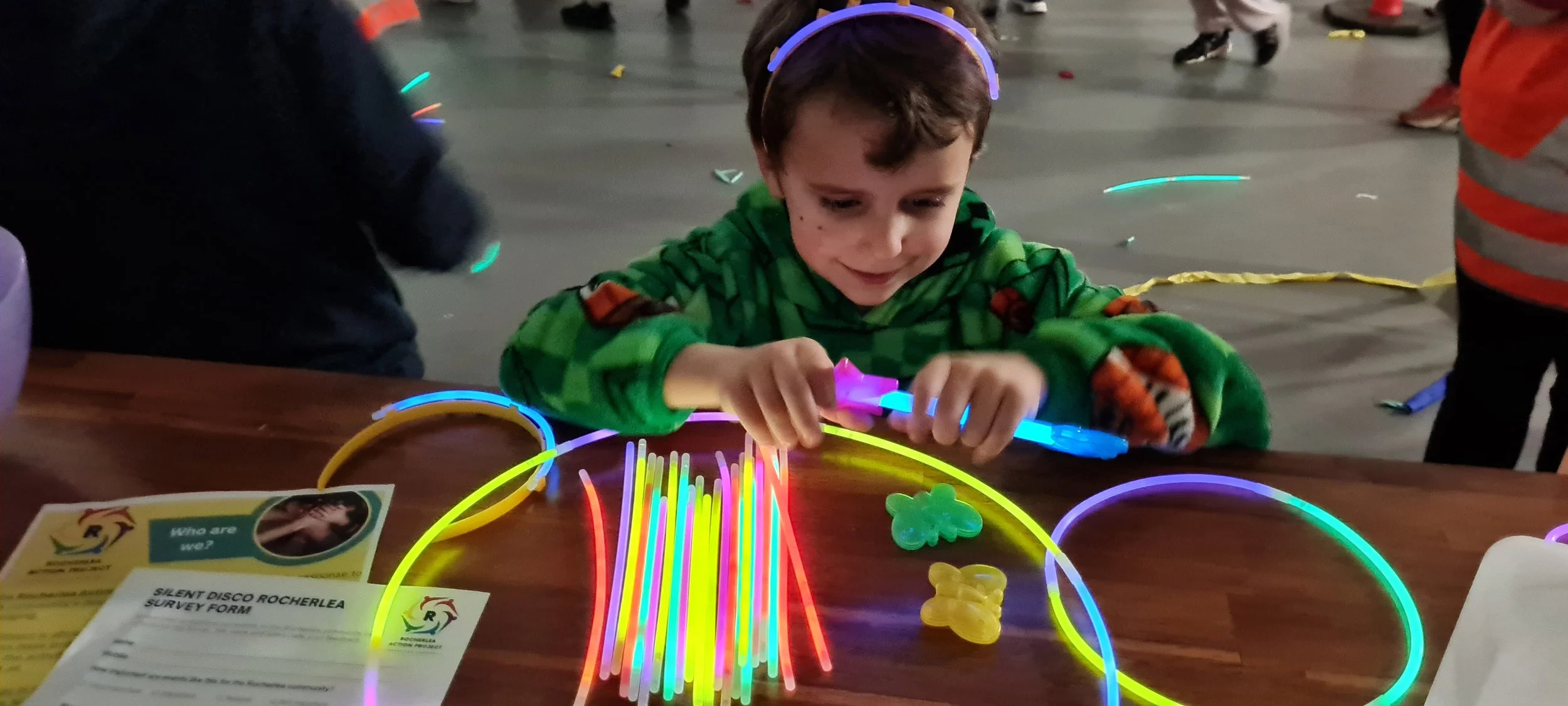A young boy with a colorful striped hoodie and a glowing headband, sitting at a table with glow sticks and gummy candies, engaged in a glow-in-the-dark activity at an indoor event.