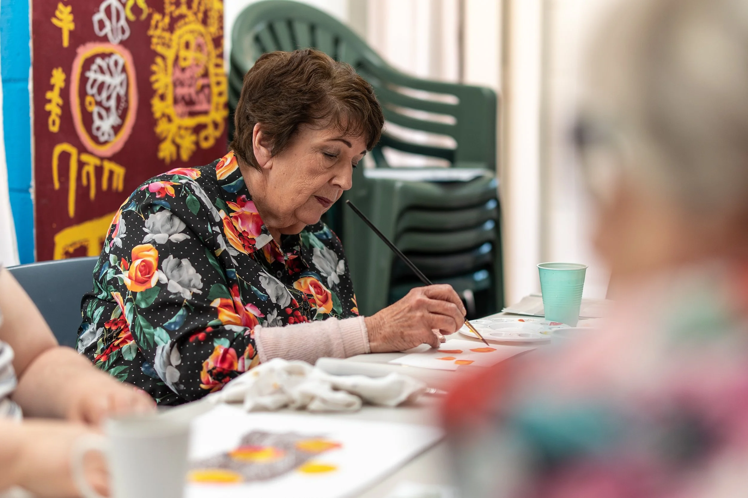 An elderly woman is sitting at a table, focused on painting a design with watercolors. She wears a black blouse with a colorful floral pattern. Behind her, purple chairs are stacked, and there is a colorful banner on the wall.