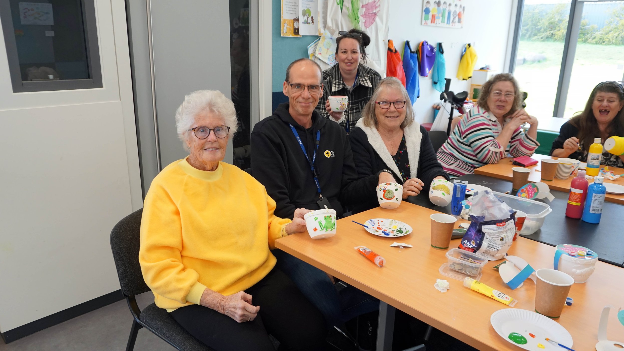 A group of six women sitting around a table in a classroom, painting and decorating ceramic mugs for a craft activity. The table has paint supplies, paper cups, and decorated mugs. Some women are smiling and enjoying the activity.