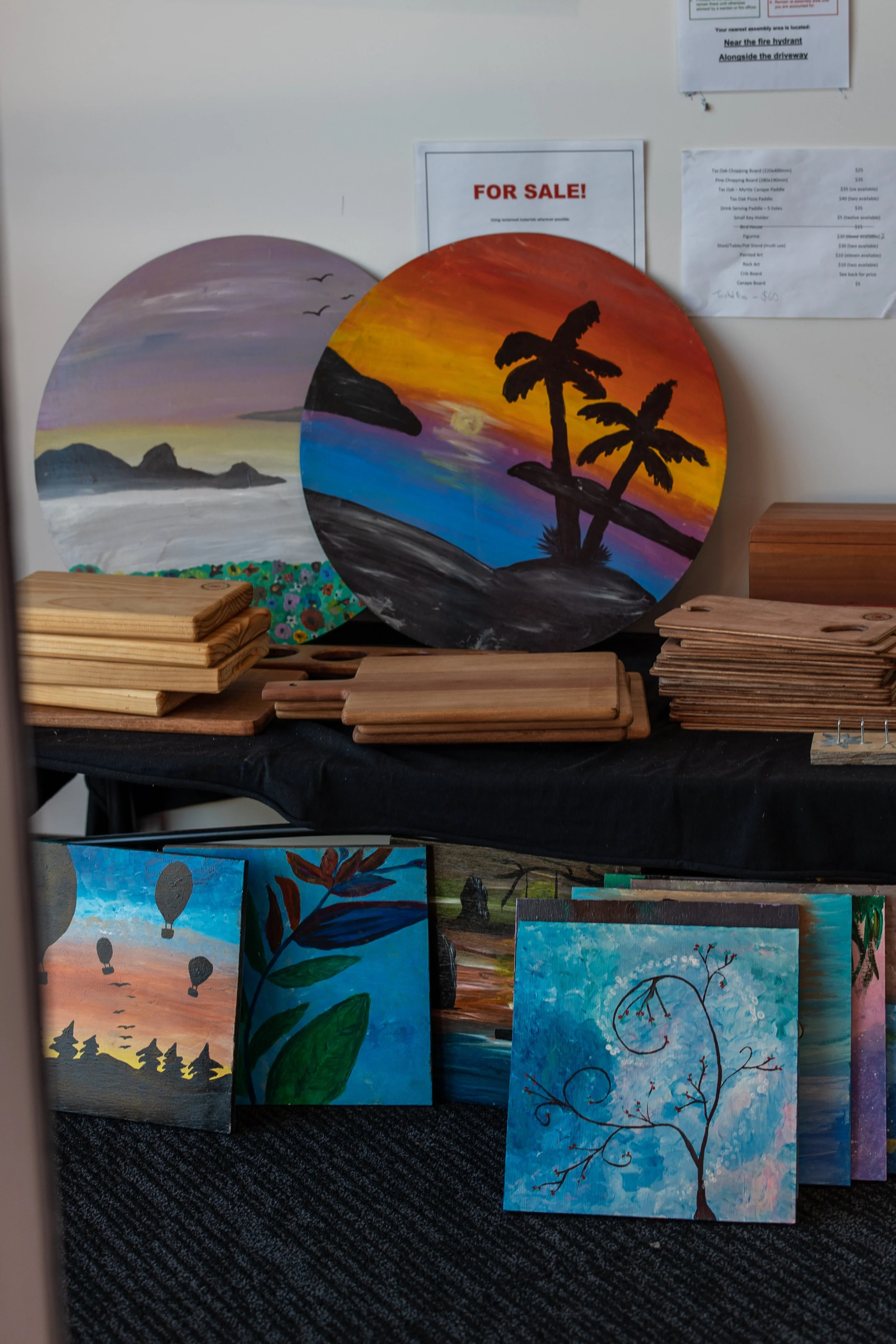 Painted circular artworks featuring landscapes and sunset scenes, along with wooden art pieces and smaller paintings of nature scenes, displayed on tables at an art sale.