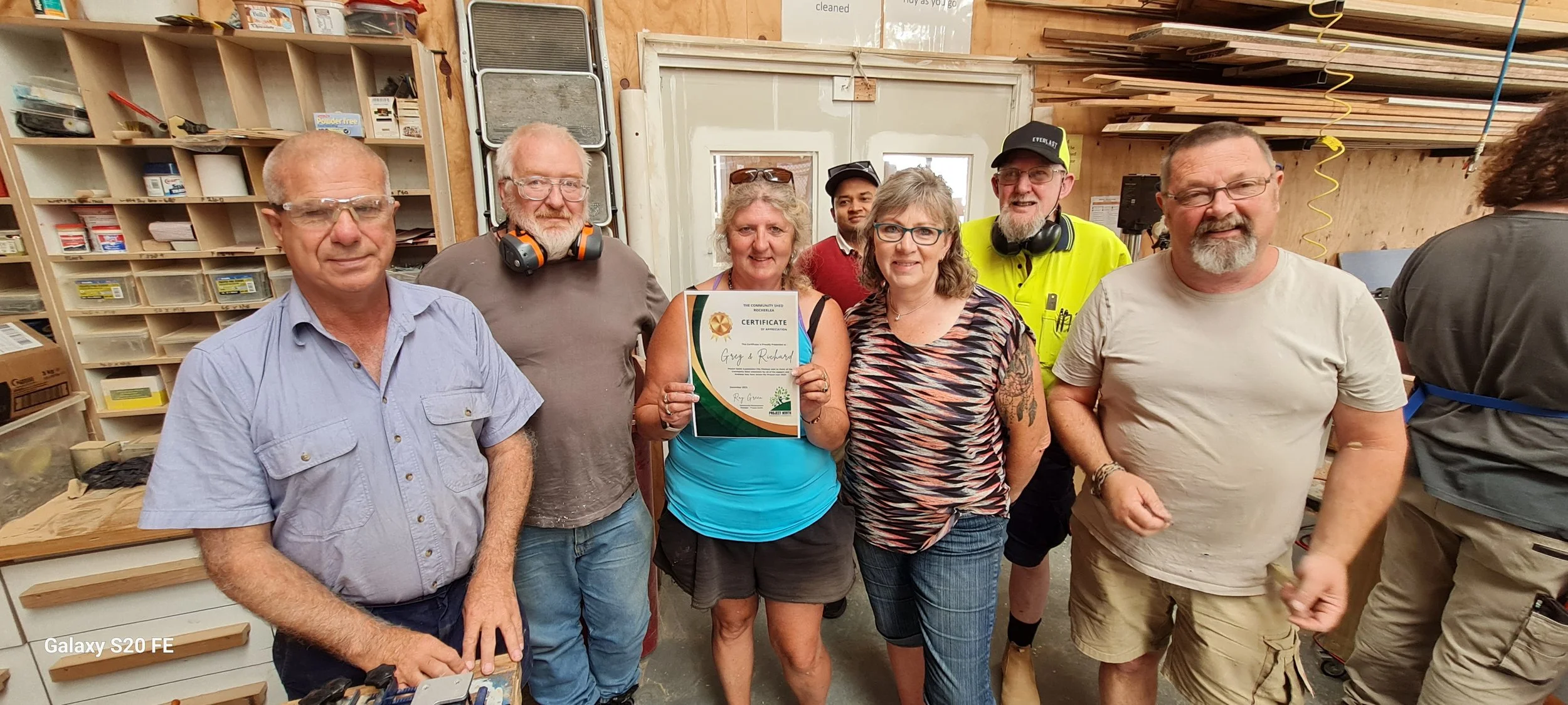 Group of six people standing in a woodworking workshop, with shelves of supplies and wood behind them. One woman in the center is holding a certificate. The individuals are smiling and appear to be celebrating an achievement.
