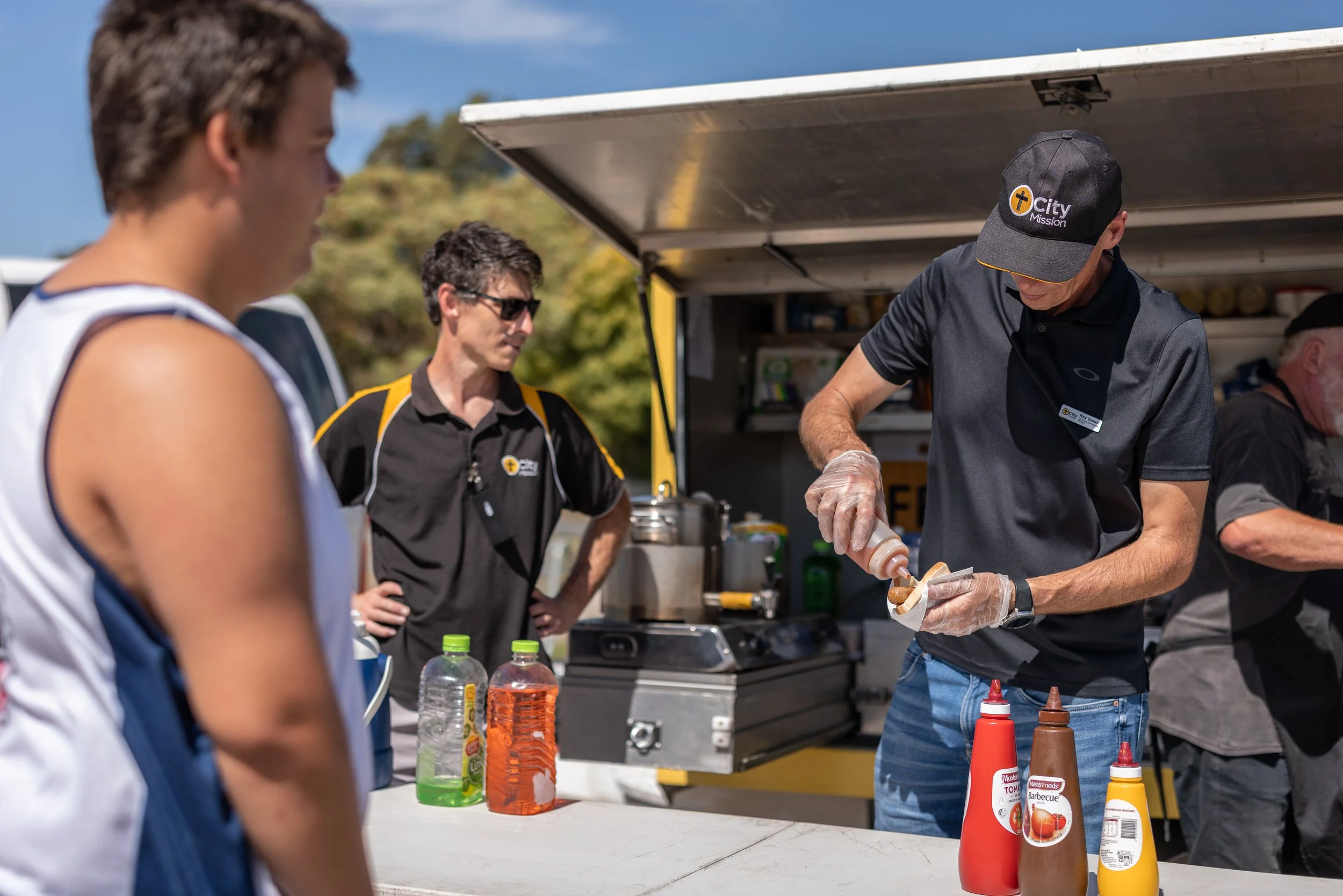 A man preparing hot dogs at a food truck, with two customers waiting in line. The man is wearing a black shirt, cap, and gloves, and is squeezing ketchup on a hot dog. There are bottles of ketchup, mustard, and barbecue sauce on the table, and two pe