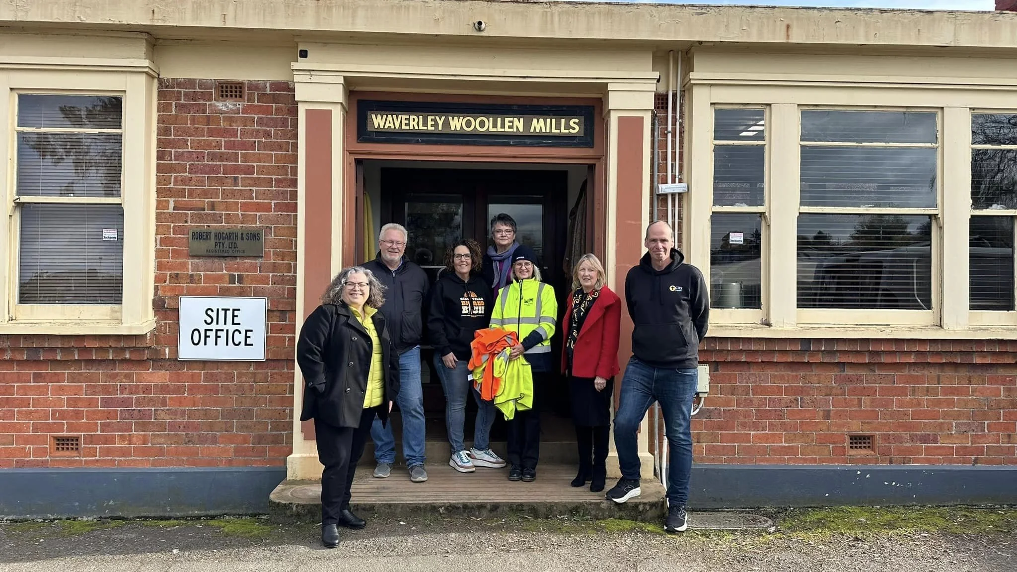 Group of seven people standing on the steps outside the Waverley Woollen Mills building, smiling and posing for the photo.
