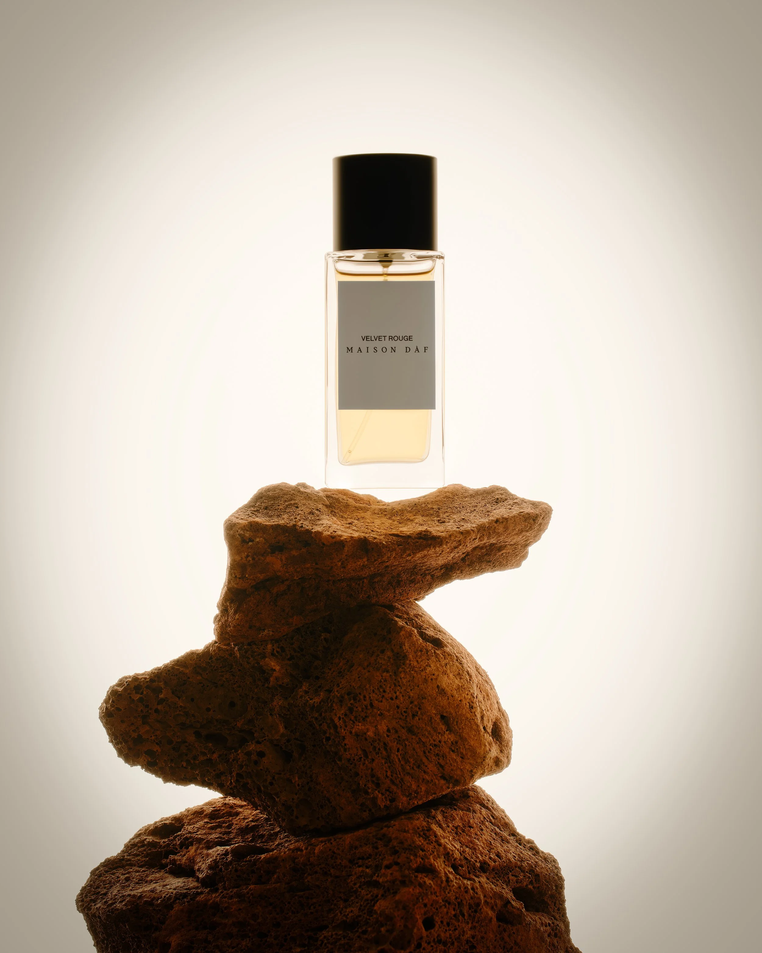 Perfume bottle labeled Velvet Rouge Maison D'Af balanced on a stack of rocks with a backlit glow.