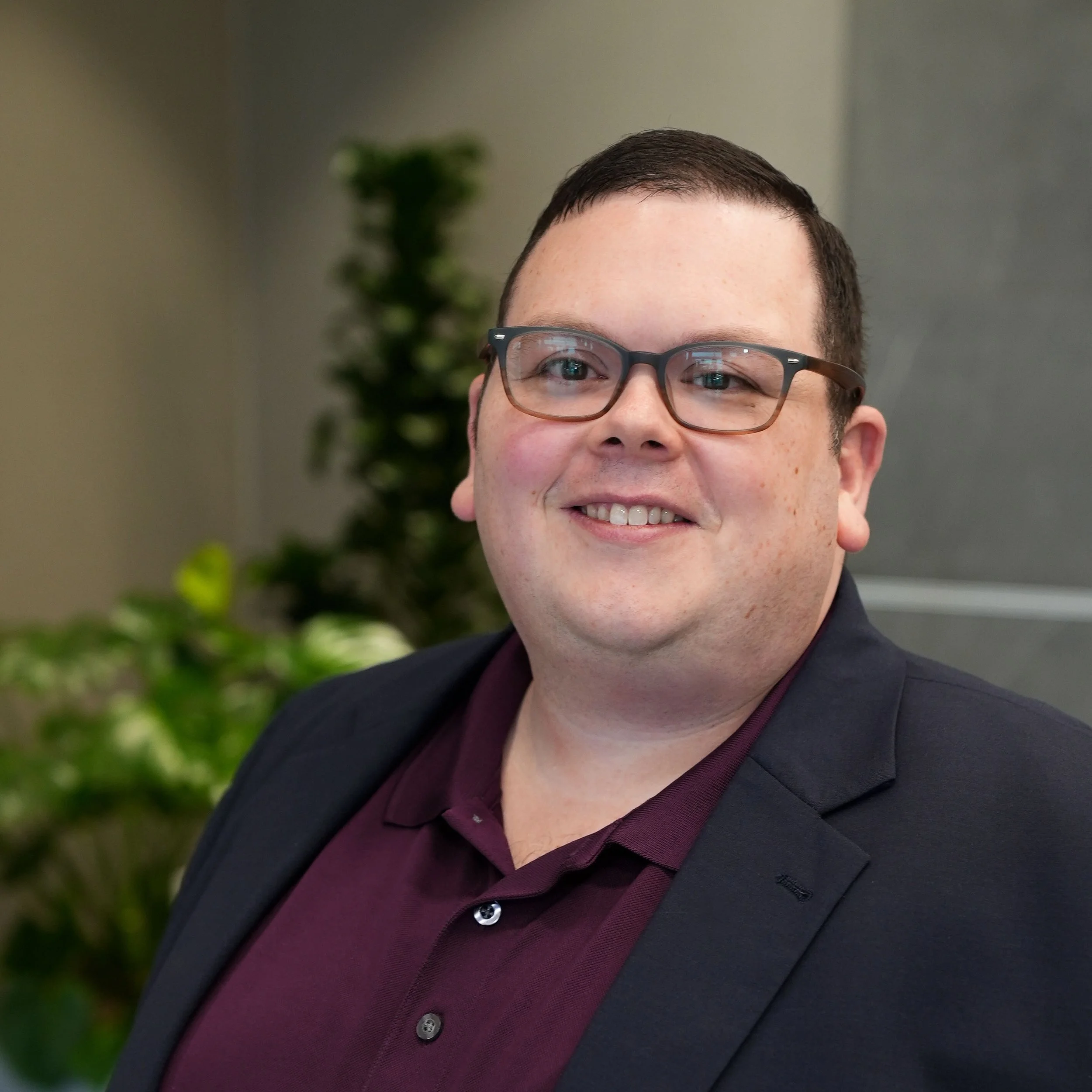 A headshot photo of Shane Jacques. He is wearing glasses, a blue sport jacket, and a plum polo shirt.