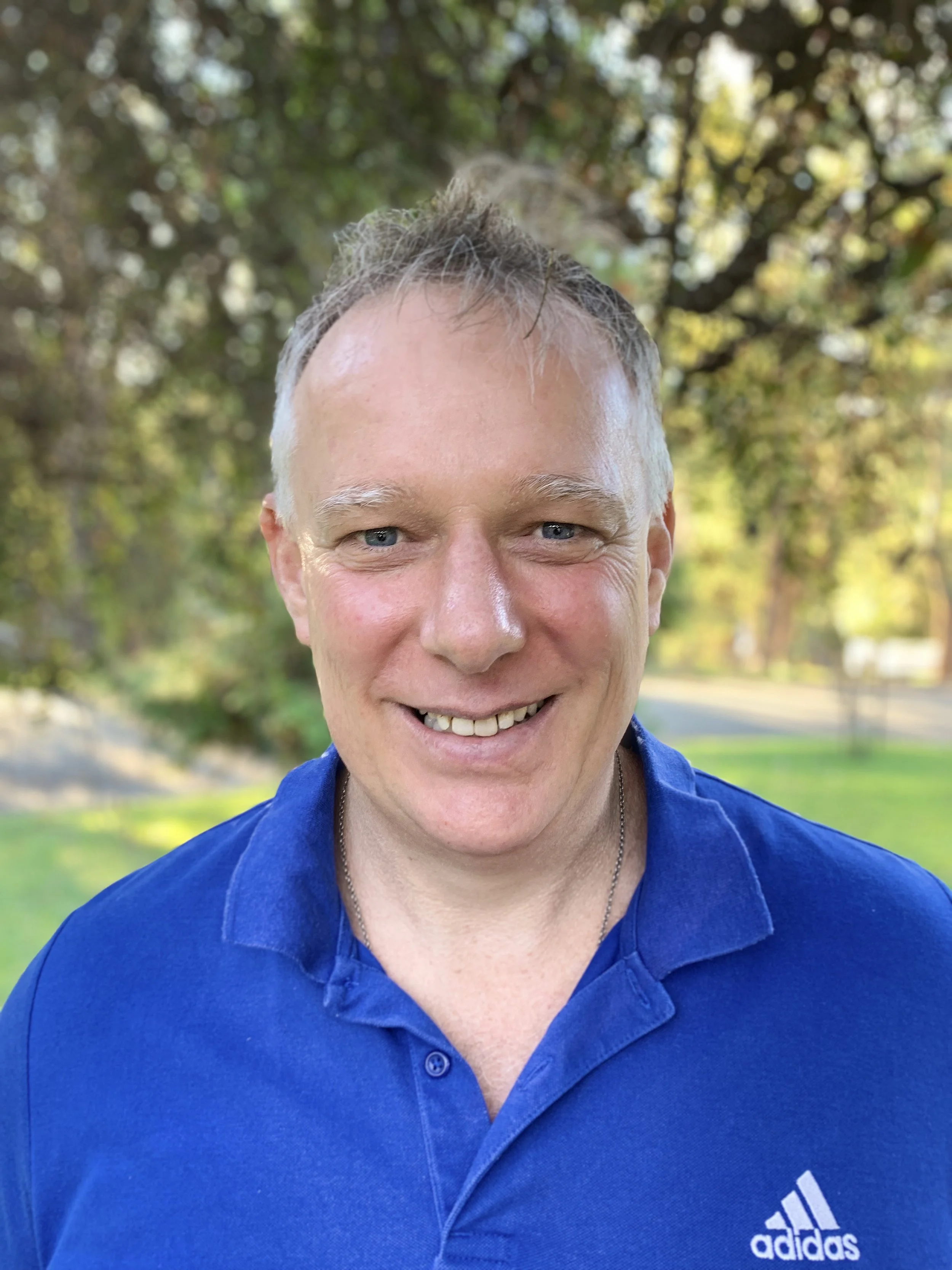 A man with short light hair, blue eyes, and a smiling face wearing a blue Adidas polo shirt outdoors with trees and sunlight in the background.