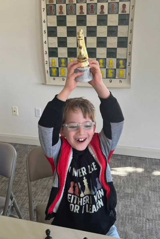 A young boy with glasses, smiling and holding a trophy above his head, sitting at a table in a room with a chessboard on the wall behind him.