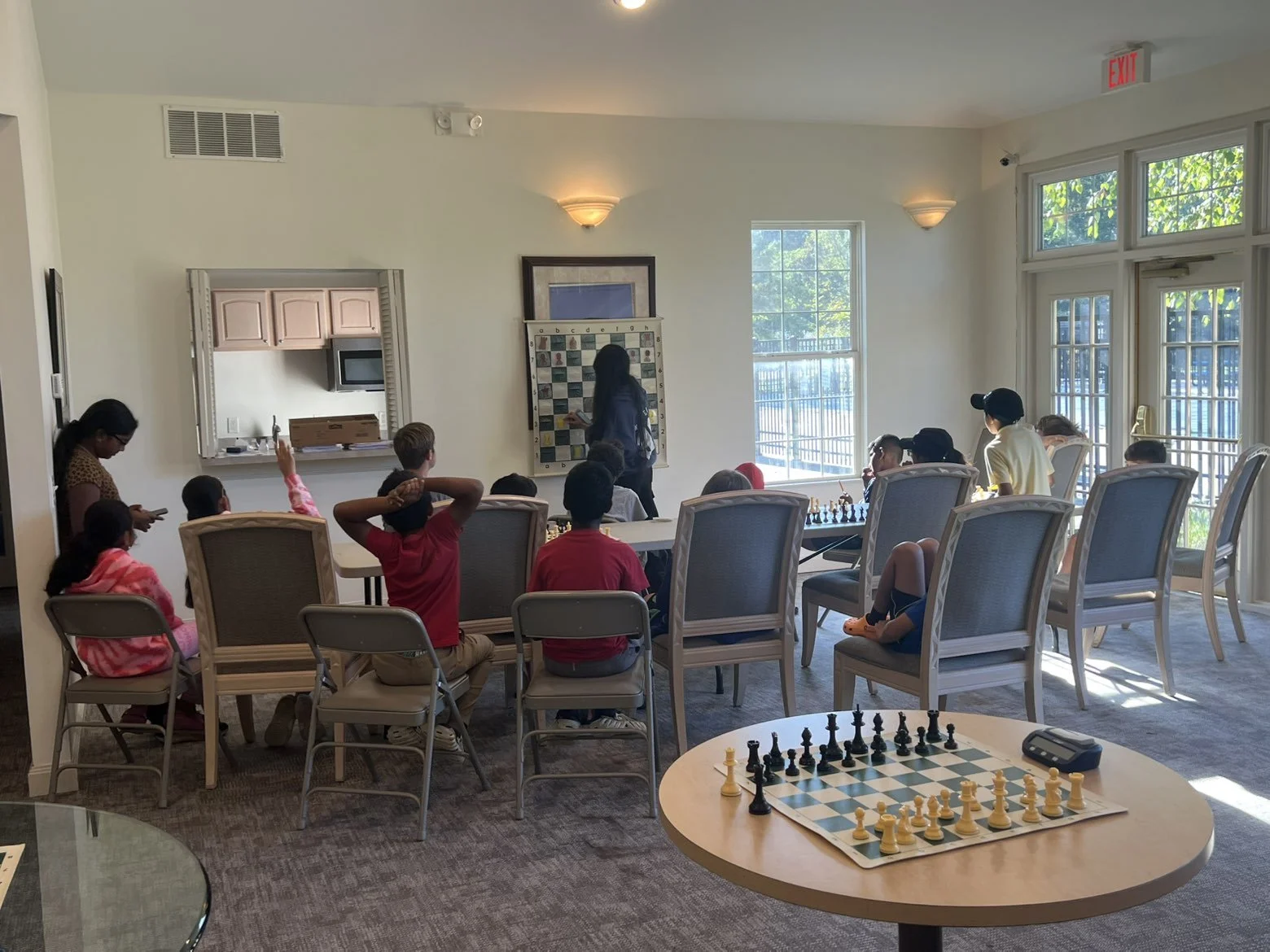 Children and adults playing chess in a well-lit room with large windows and an open kitchen window. A chessboard with pieces set up is on a round table in the foreground.