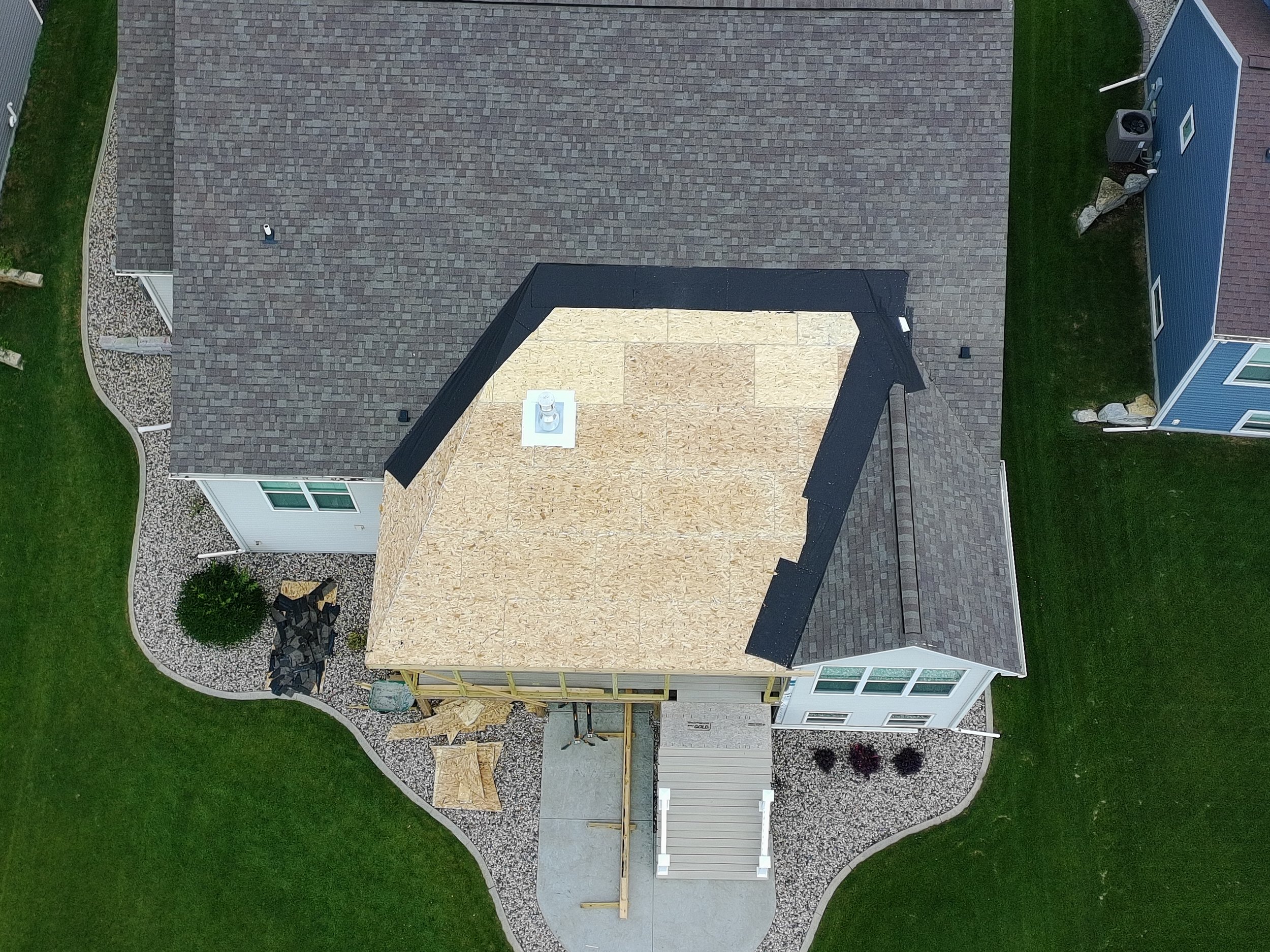 Aerial view of a house under construction with a new roof in progress, showing a partially shingled roof and plywood decking, surrounded by a landscaped yard with gravel and grass.