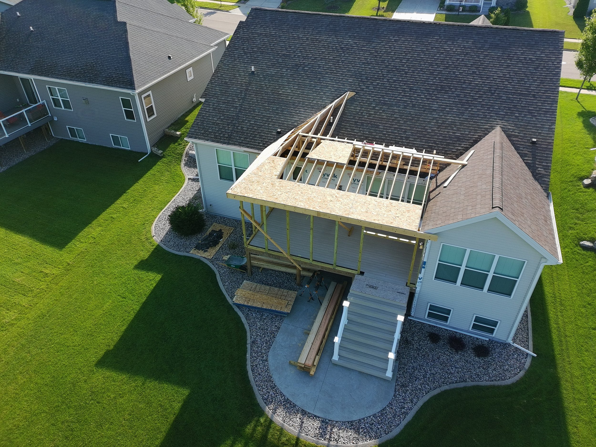 An aerial view of a house under construction, showing a partially built wooden balcony or deck extension above the front entrance, with construction materials and tools visible on the site. The house has white siding, large windows, and a shingled ro