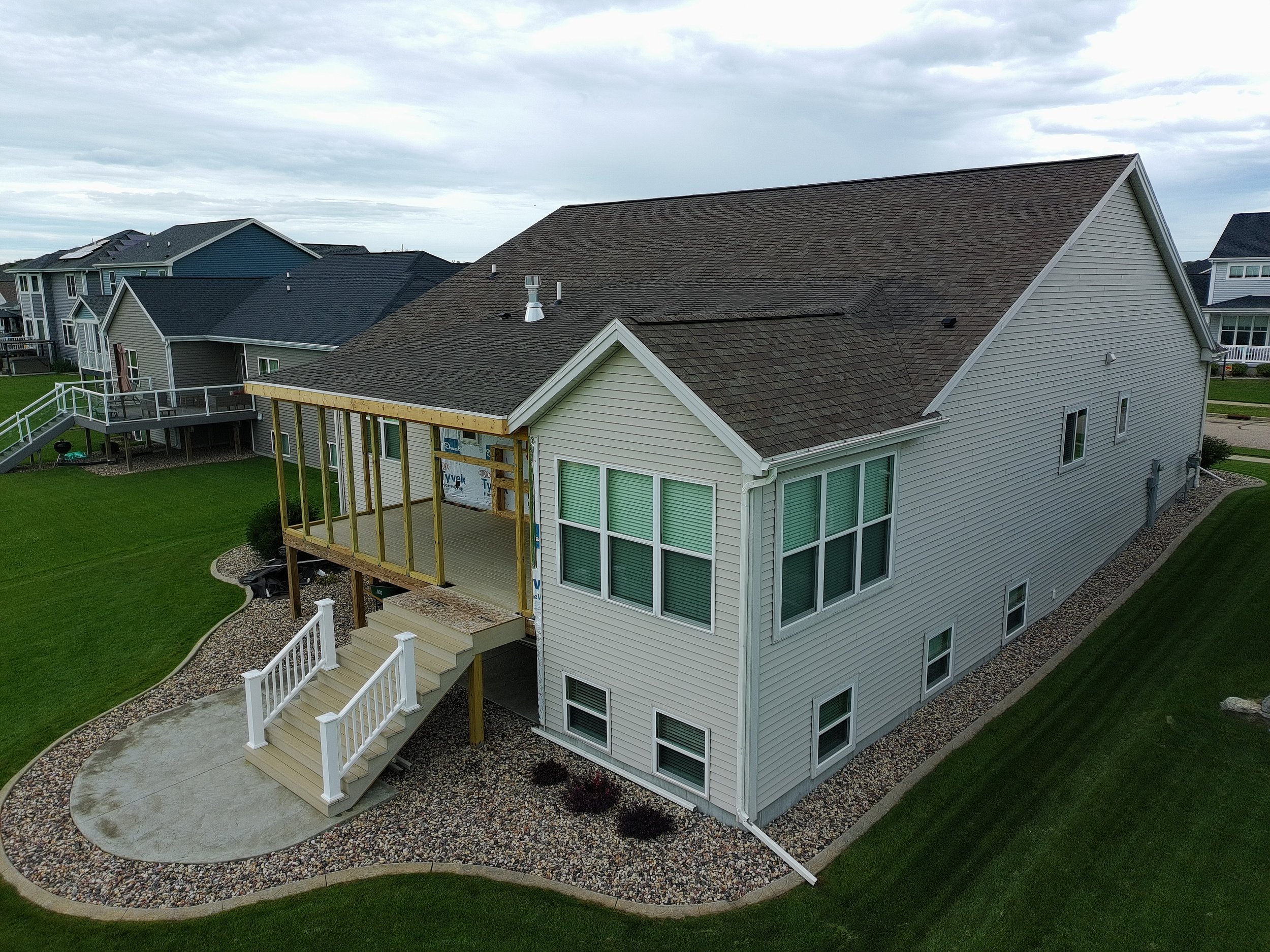A two-story house with vinyl siding and several windows, with an elevated deck under construction on the back, supported by wooden posts with stairs leading down to a stone patio surrounded by a landscaped yard.
