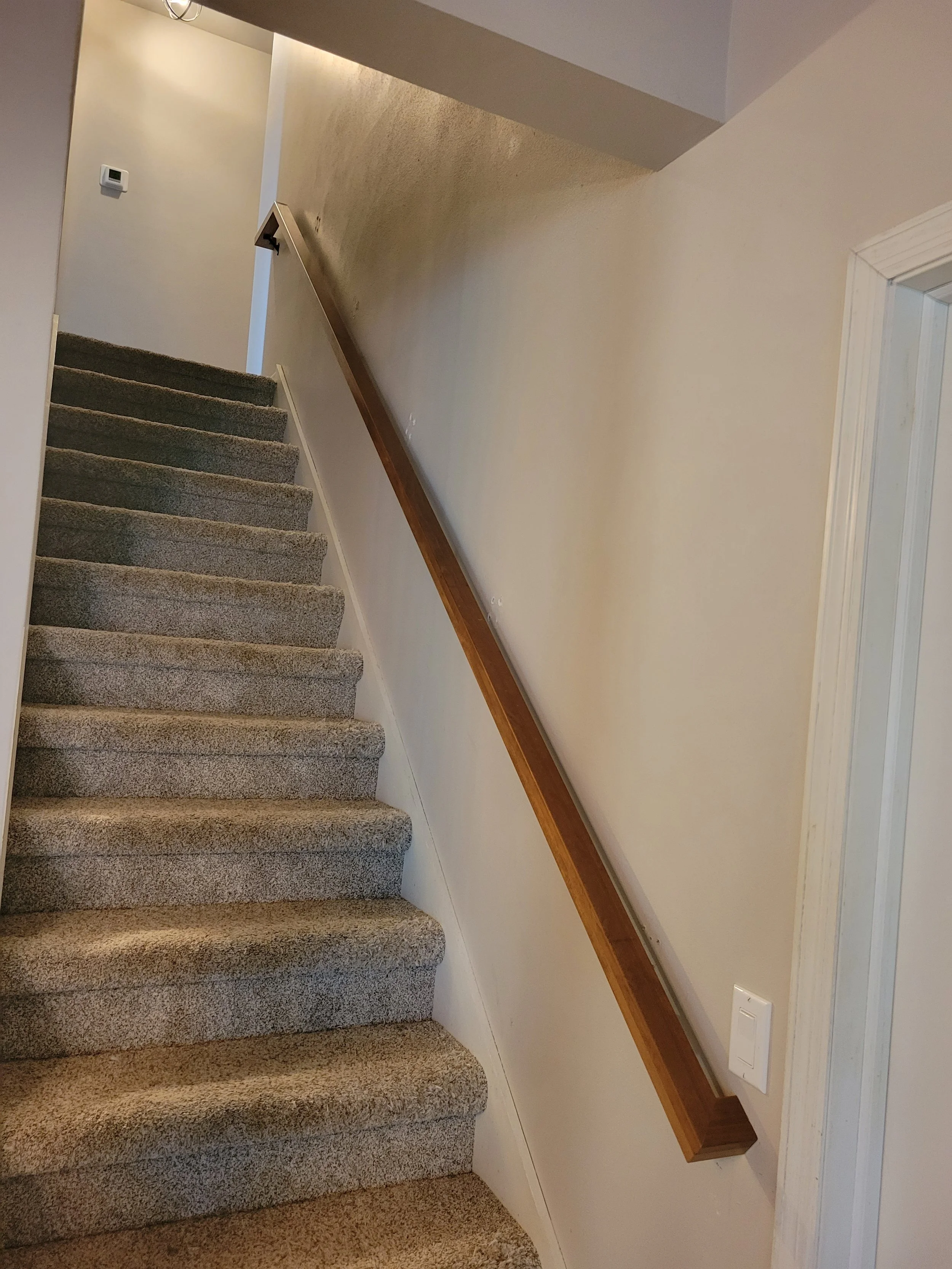 Photo of a staircase with gray carpeted steps, a wooden handrail on the right side, a beige wall, and a light switch near the bottom right corner.