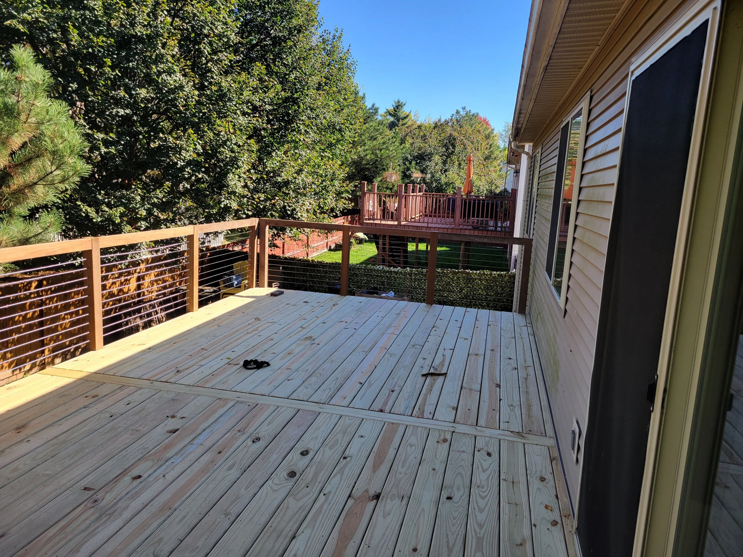 Newly constructed wooden deck attached to a house with beige siding, overlooking a backyard with a larger elevated deck and a picket fence, surrounded by trees and under a clear blue sky.