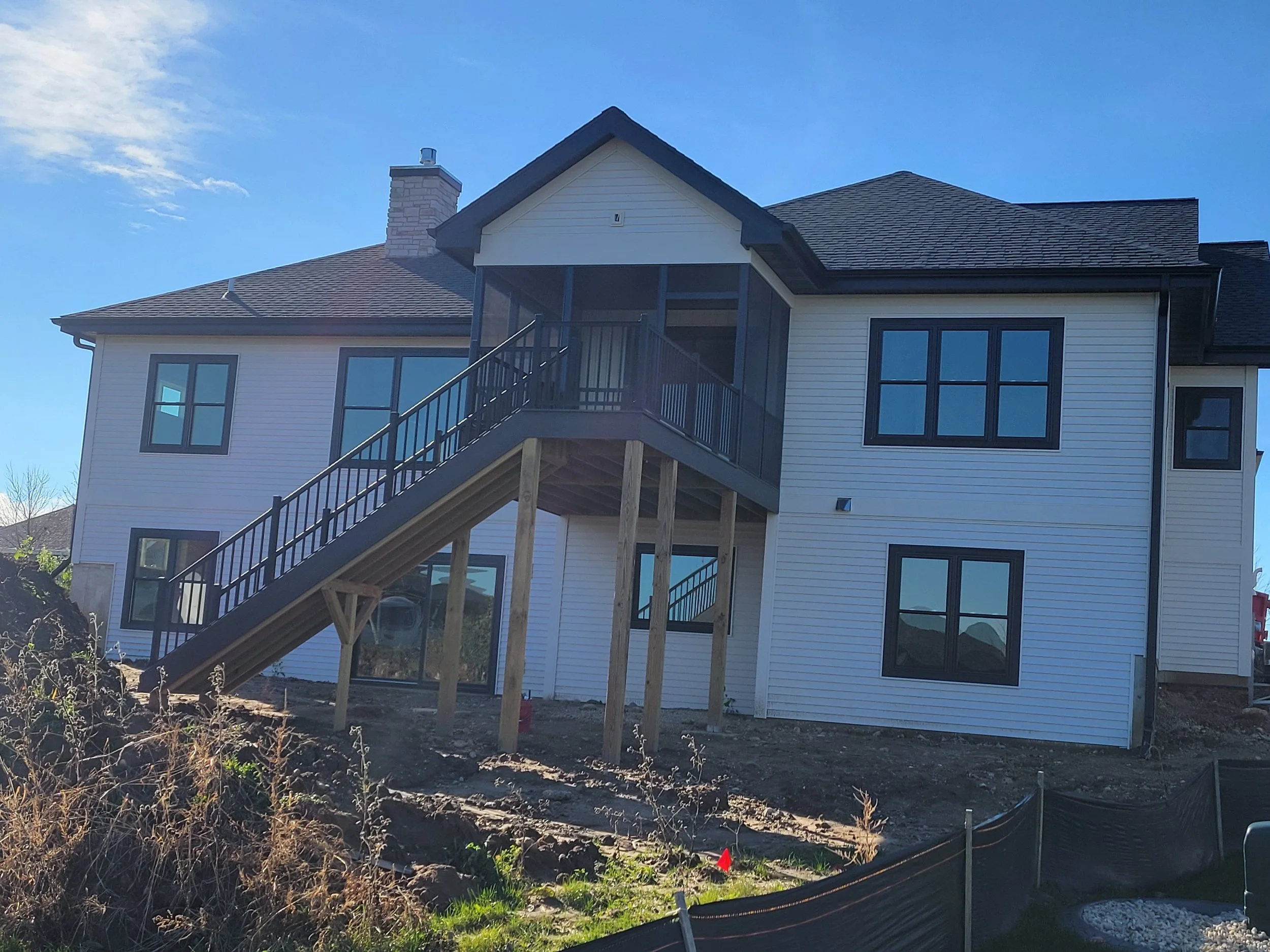 New two-story house with white siding, black-framed windows, and an external staircase leading to a porch on the upper level, under construction with exposed dirt ground.
