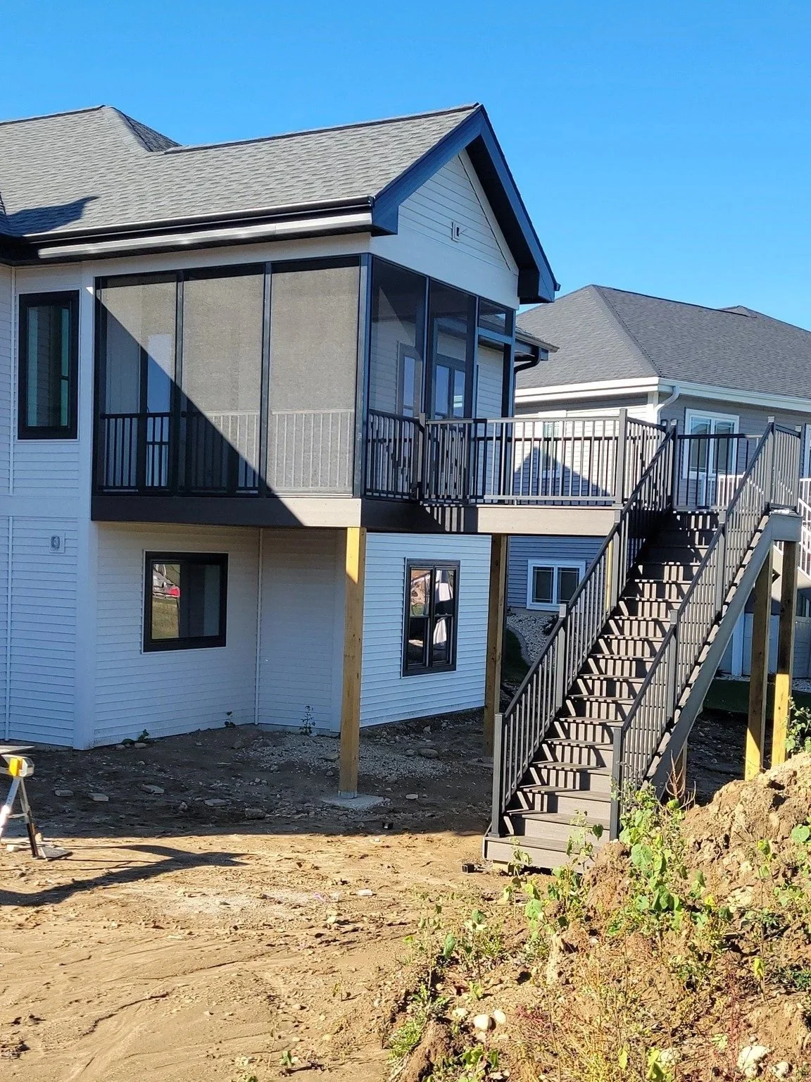 Newly constructed two-story house with white siding, black accents, and an unfinished yard. The house has a screened porch on the upper level and stairs leading down to the ground.