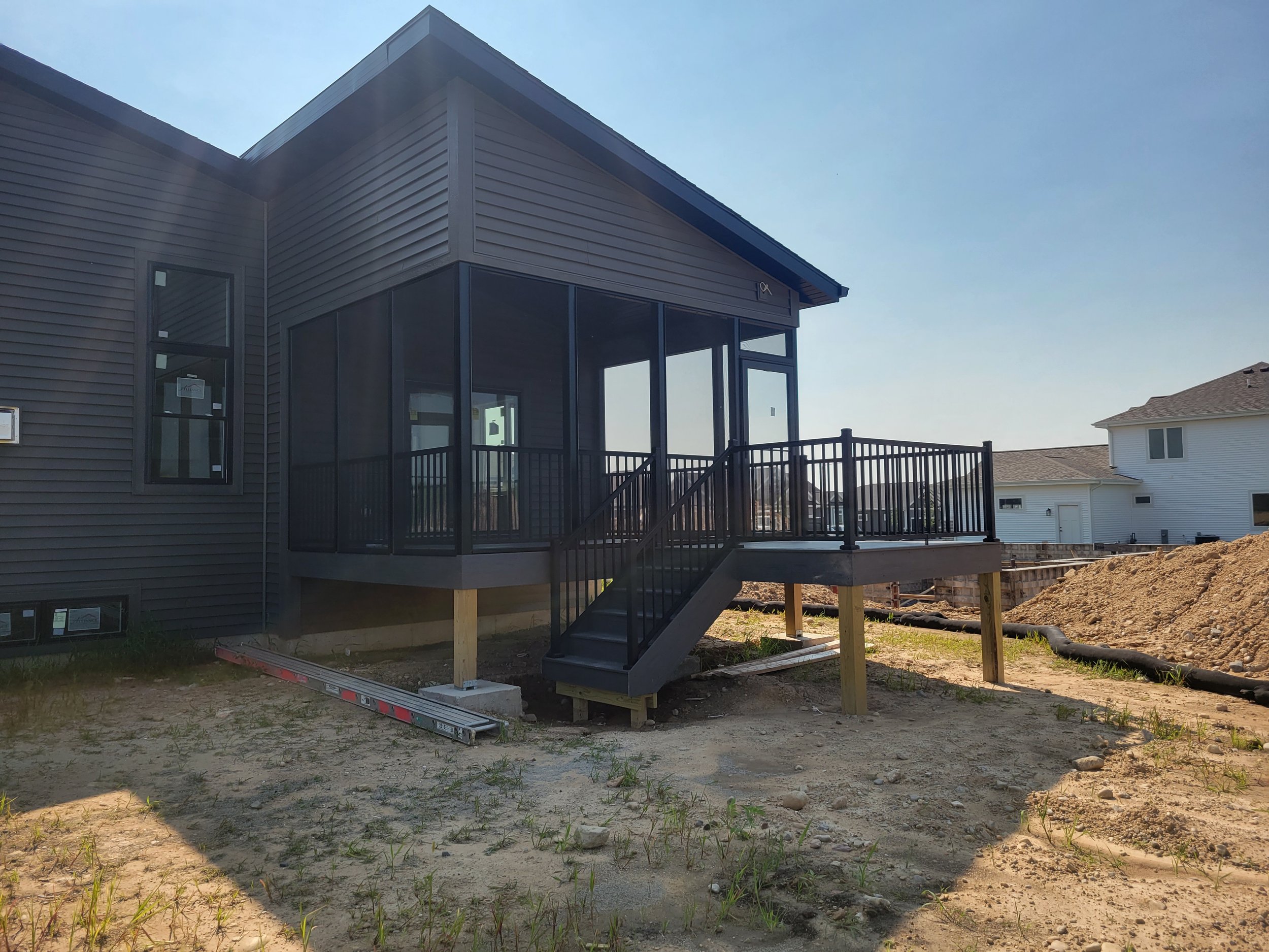 A new house with a black front porch and railing, located in a construction site with dirt and grass, with neighboring houses visible in the background.
