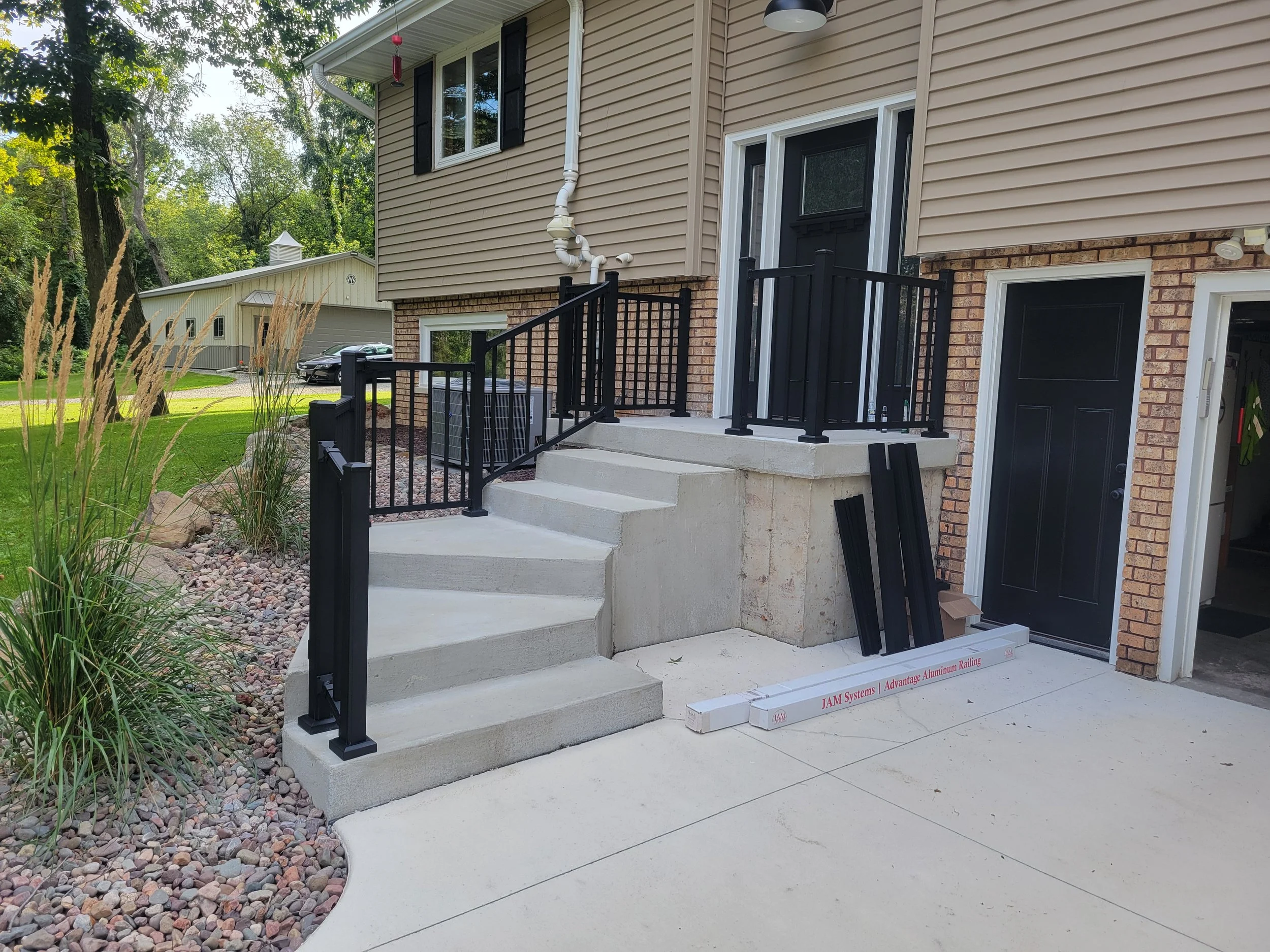 Newly installed black metal railing on concrete stairs outside a house with brick and siding exterior, with landscaping on the side.