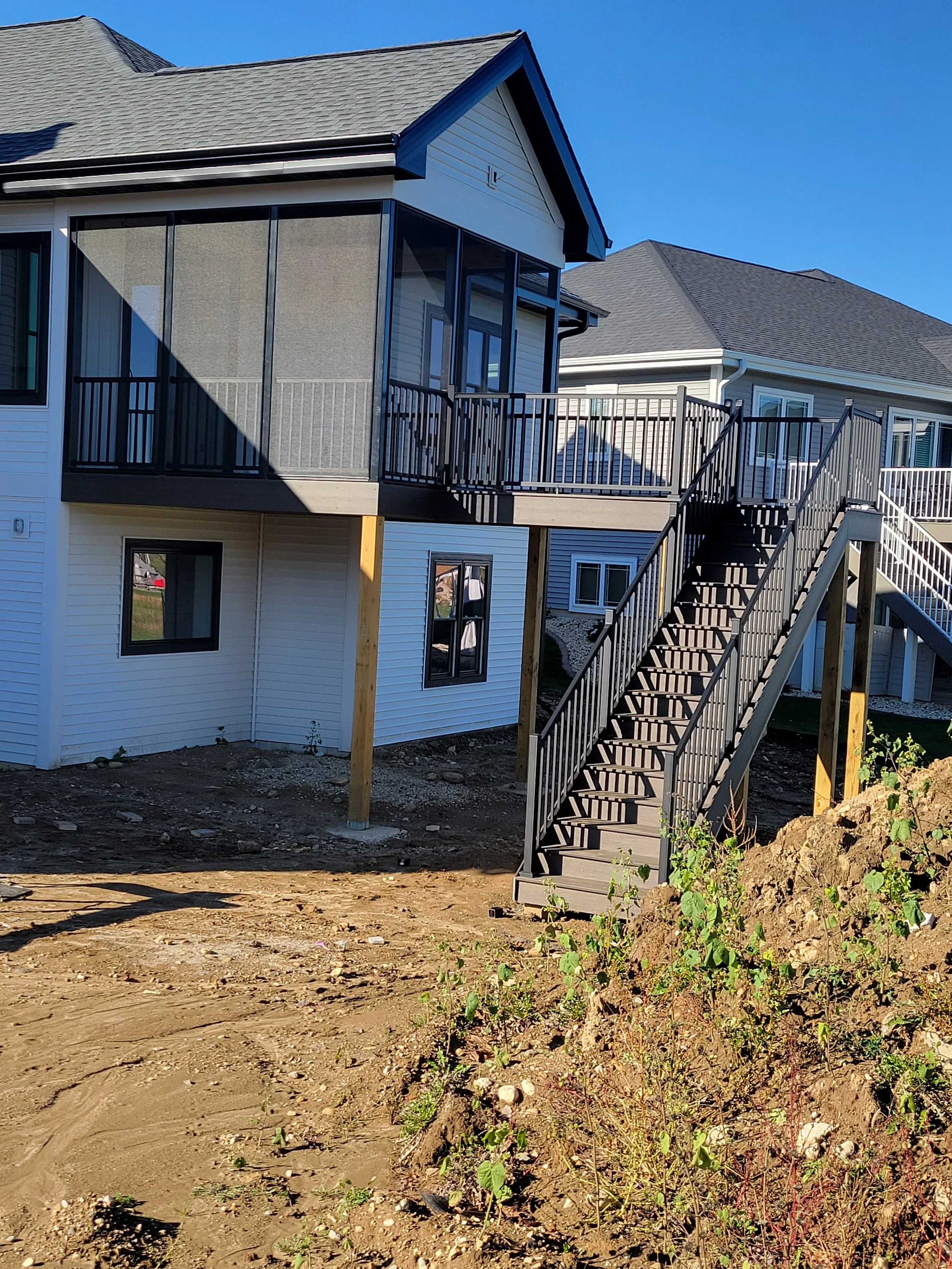The photo shows the backyard of a newly constructed house with a wooden deck and stairs. The house has white siding with black-framed windows, and the deck is supported by wooden posts. The area below the deck is unfinished, with exposed dirt and sma