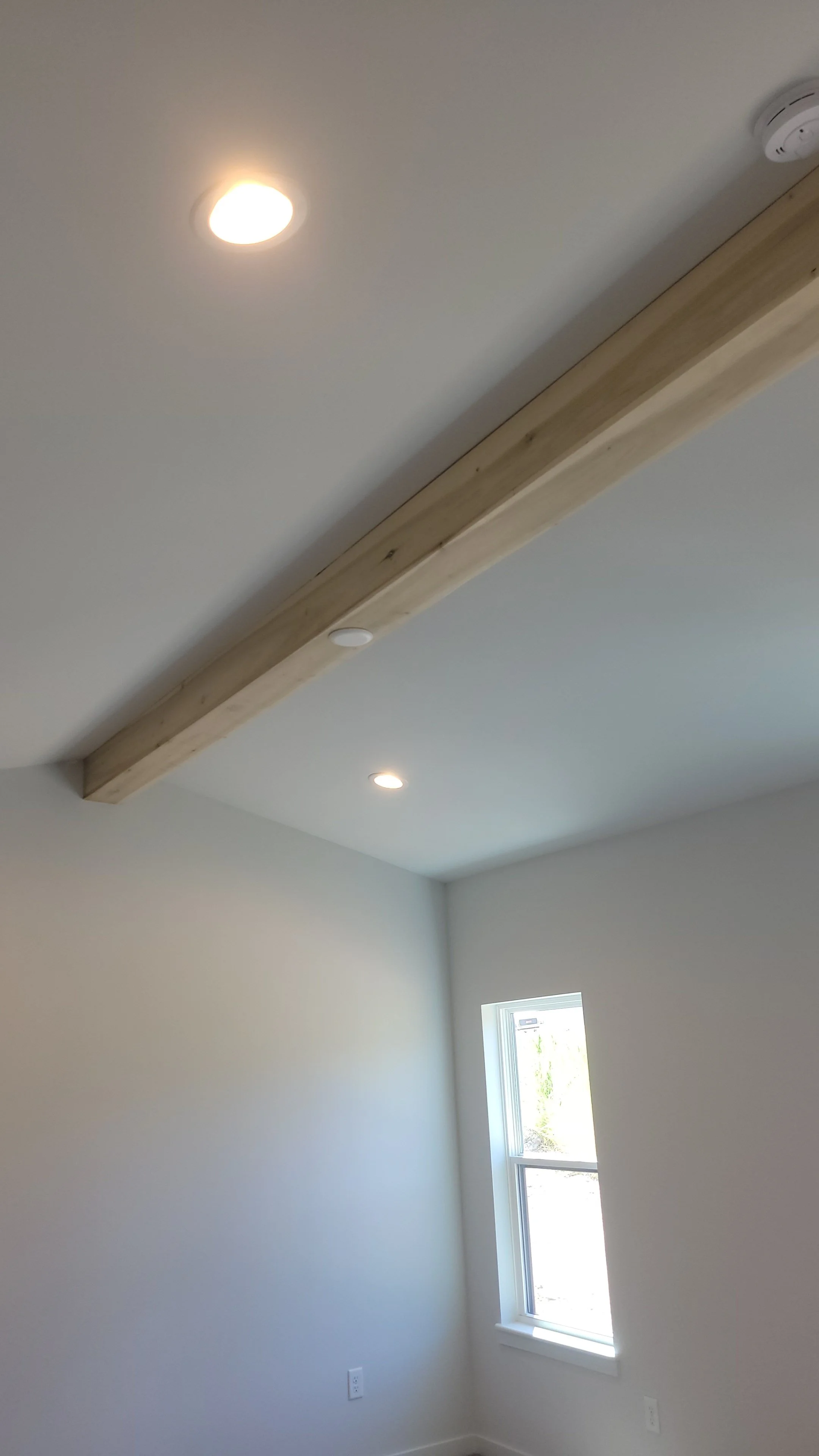 Interior of a room with white walls, a small window, and a ceiling with recessed lighting and a wooden beam.