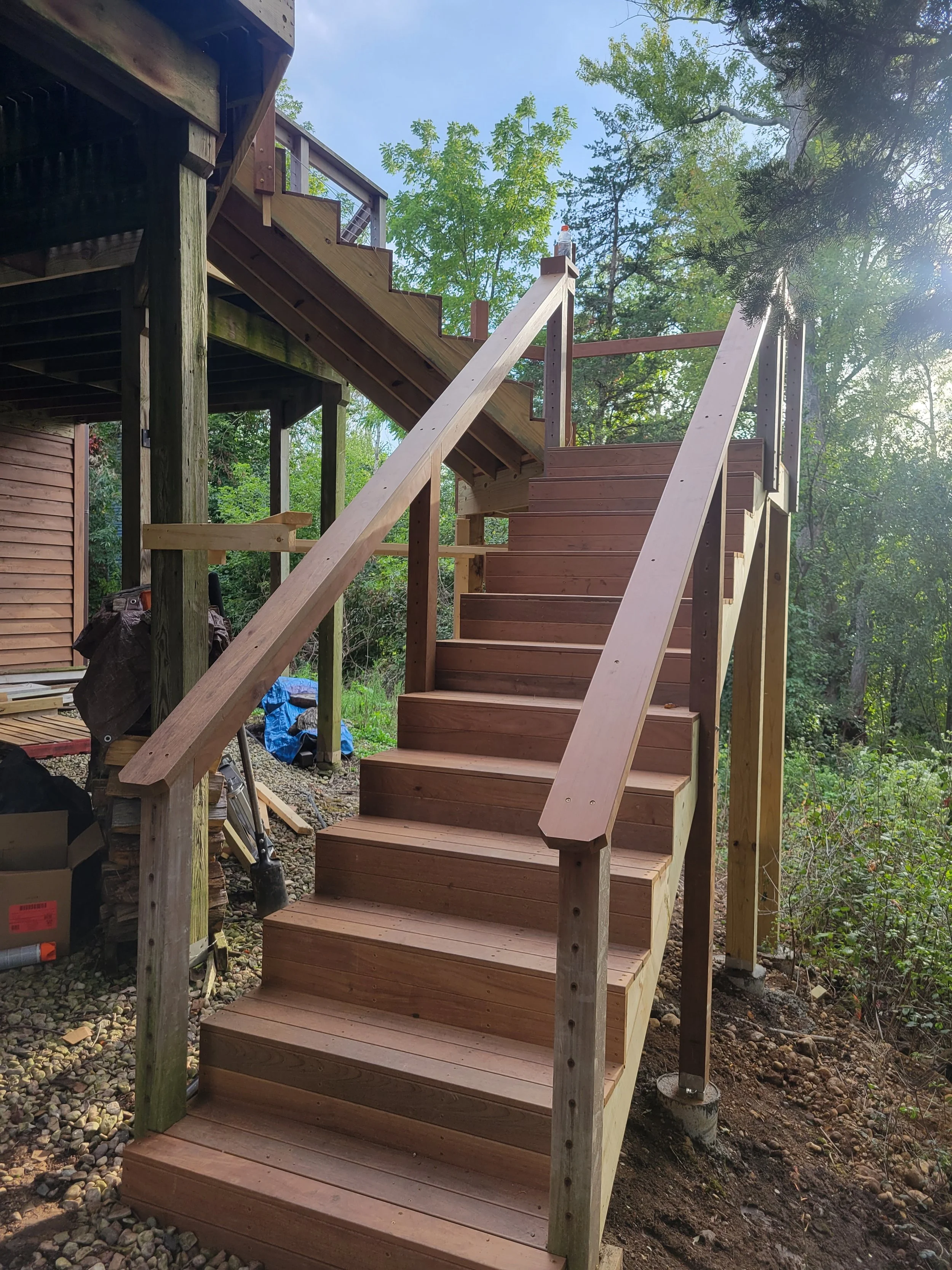 Wooden outdoor staircase leading up to a deck, surrounded by trees and construction materials on the ground.