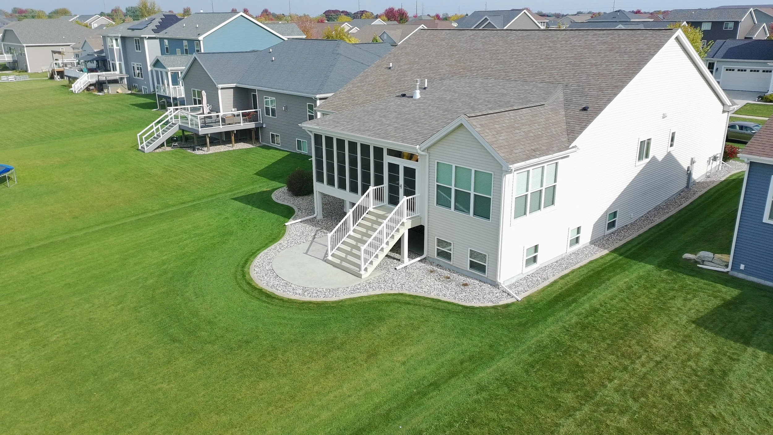 An aerial view of a suburban neighborhood showing multiple houses with well-maintained green lawns. The focus is on a large white house with a screened porch and staircase leading to the backyard, featuring a gravel pathway around it. Other houses wi