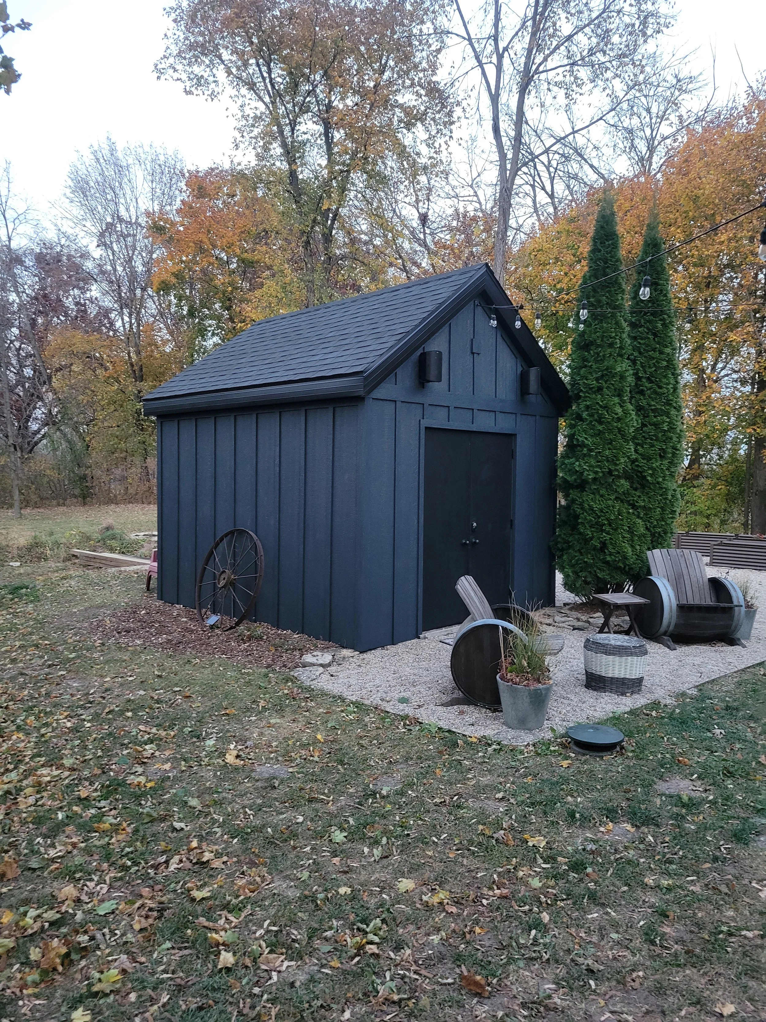 A small black shed with a sloped roof, surrounded by outdoor furniture and decorations, with fall trees in the background.