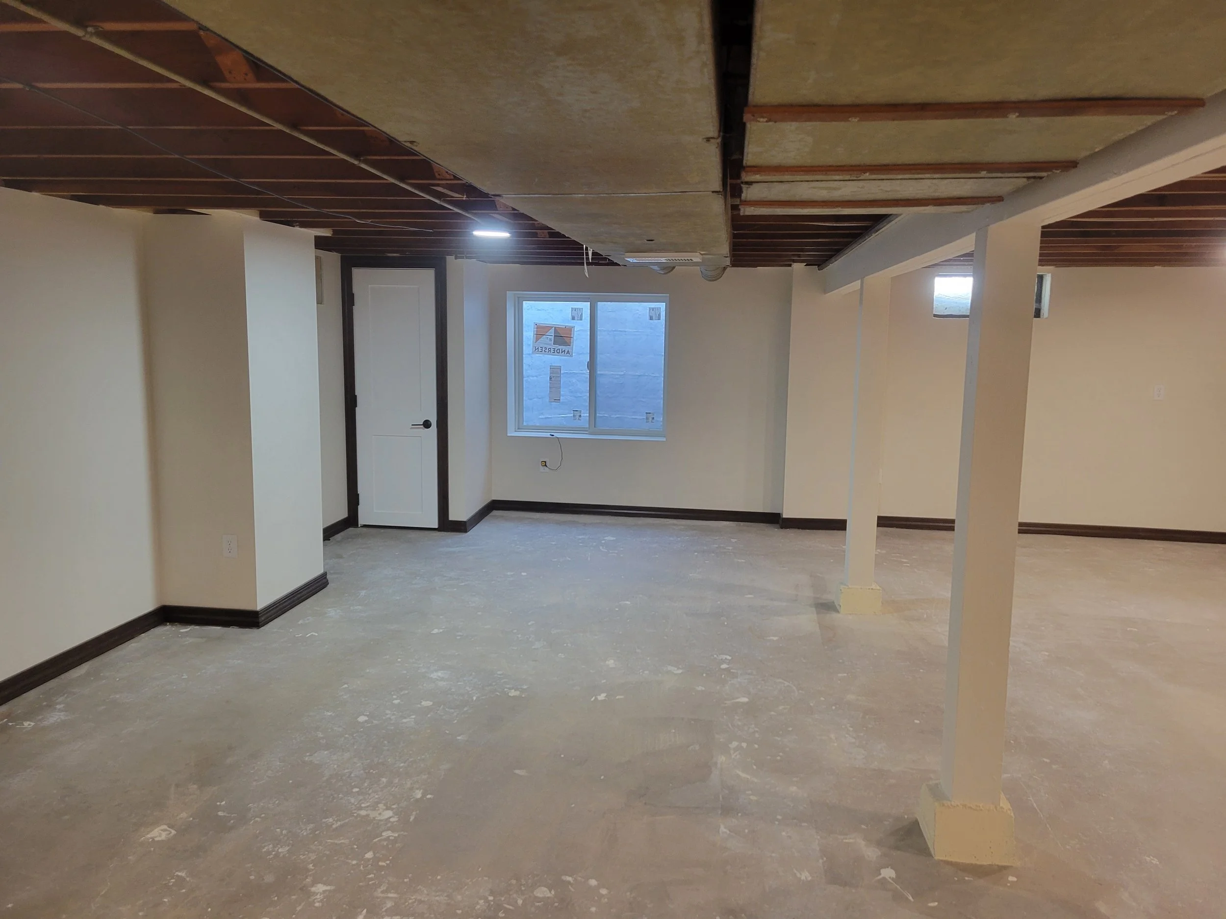 Empty room under construction with exposed beams, a window, and partially finished floor.