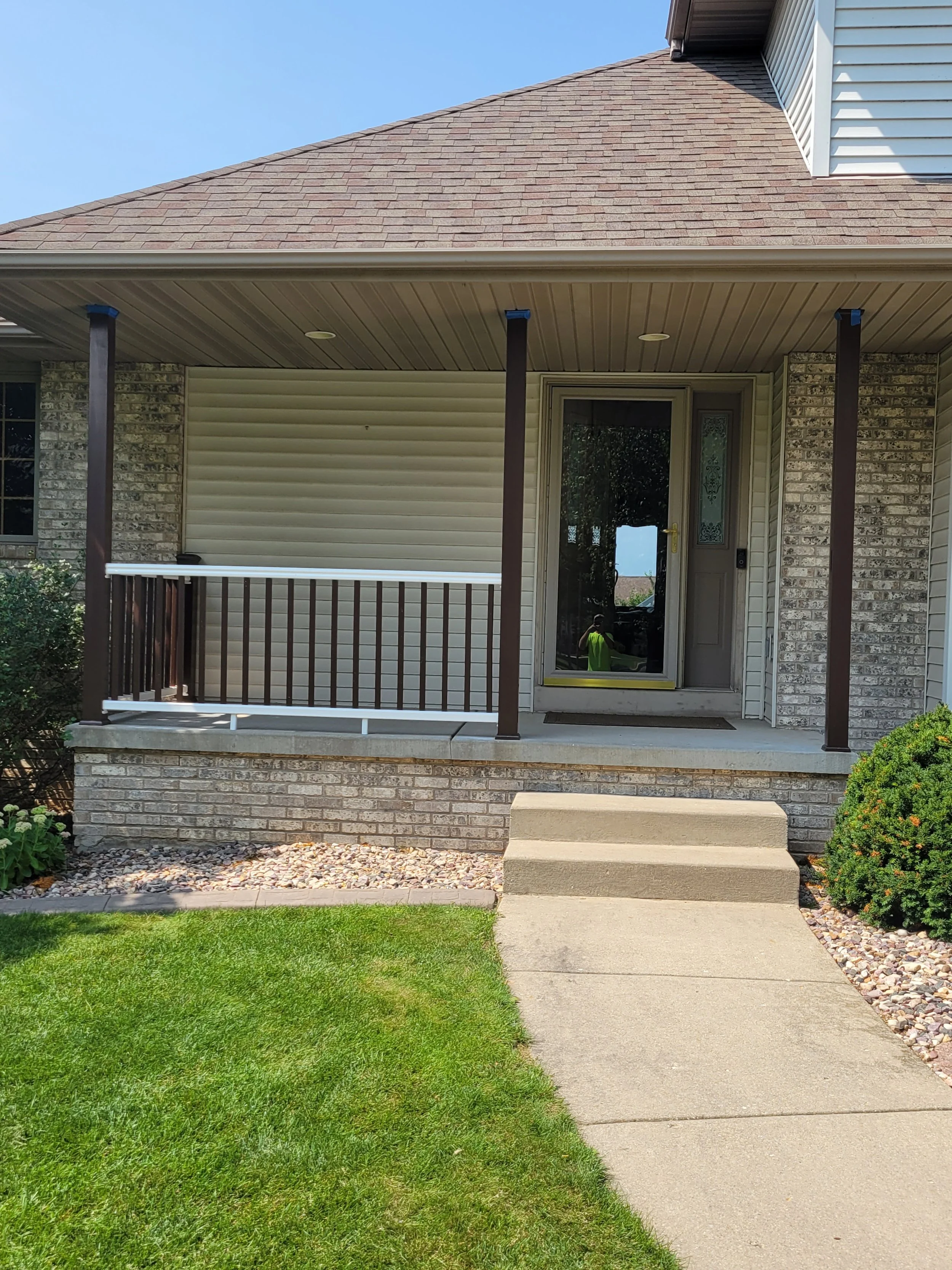 Front porch of a house with a concrete pathway, small steps, a grassy lawn, and a shrub on the right. The porch has a brown railing, three dark brown columns, and a front door with a glass panel.