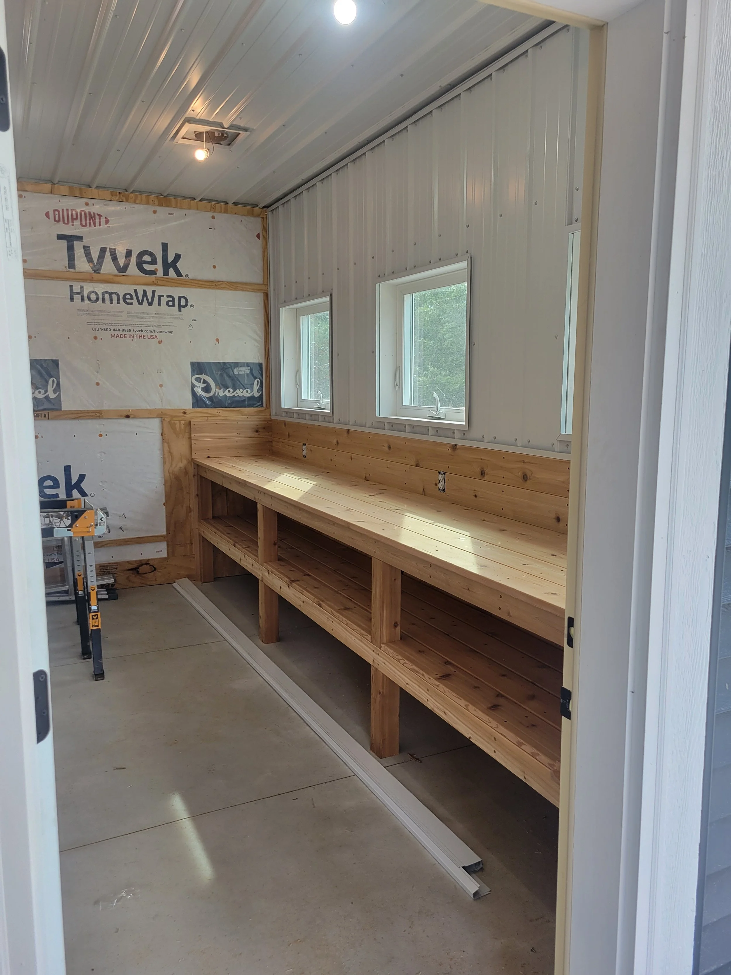 Inside a workshop or garage with a long wooden workbench against the wall beneath three small windows, power outlets, and a light fixture on the ceiling. The wall behind the workbench shows insulation with Tyvek HomeWrap branding.