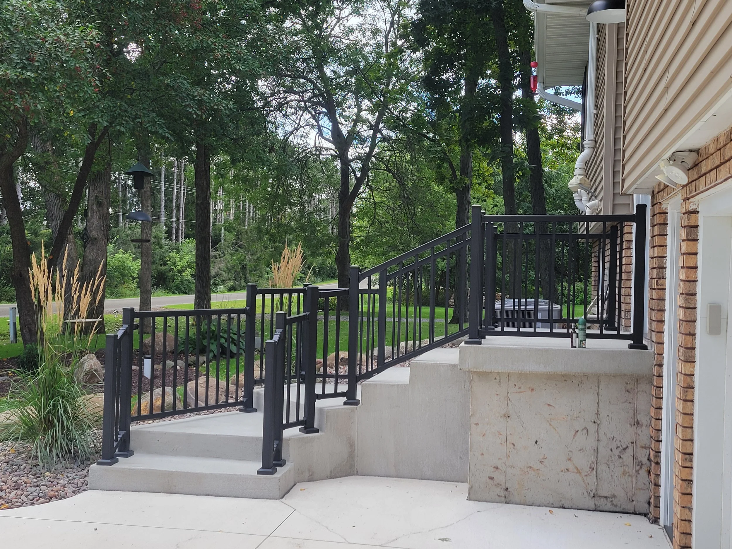 Outside view of a house with a concrete ramp and stairs, black metal railings, surrounded by trees and landscaping.