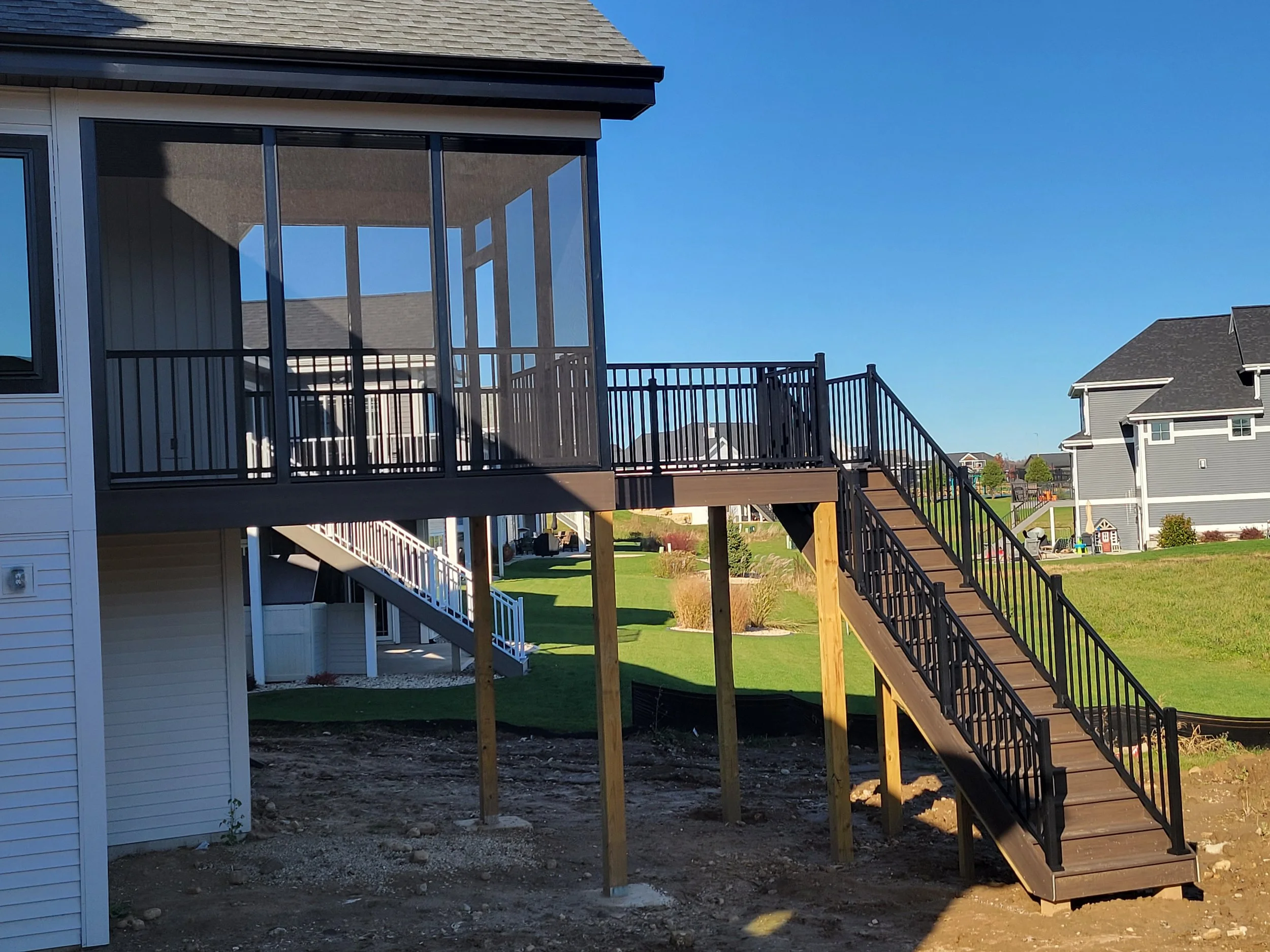 A house's elevated deck with black metal railing and wooden stairs leading down to the yard, supported by wooden posts, in a residential neighborhood with houses and green lawns.