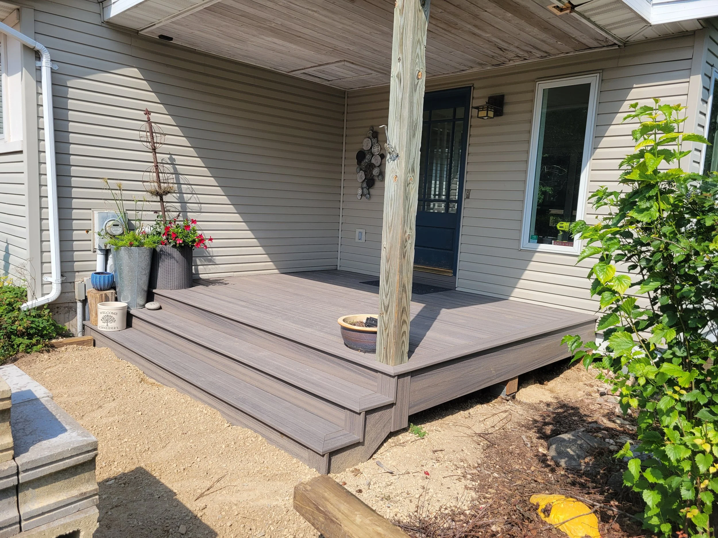 A small elevated porch with wood decking and three steps leading up to to the house, with various potted plants and gardening items on it, and a house with a door and window in the background.