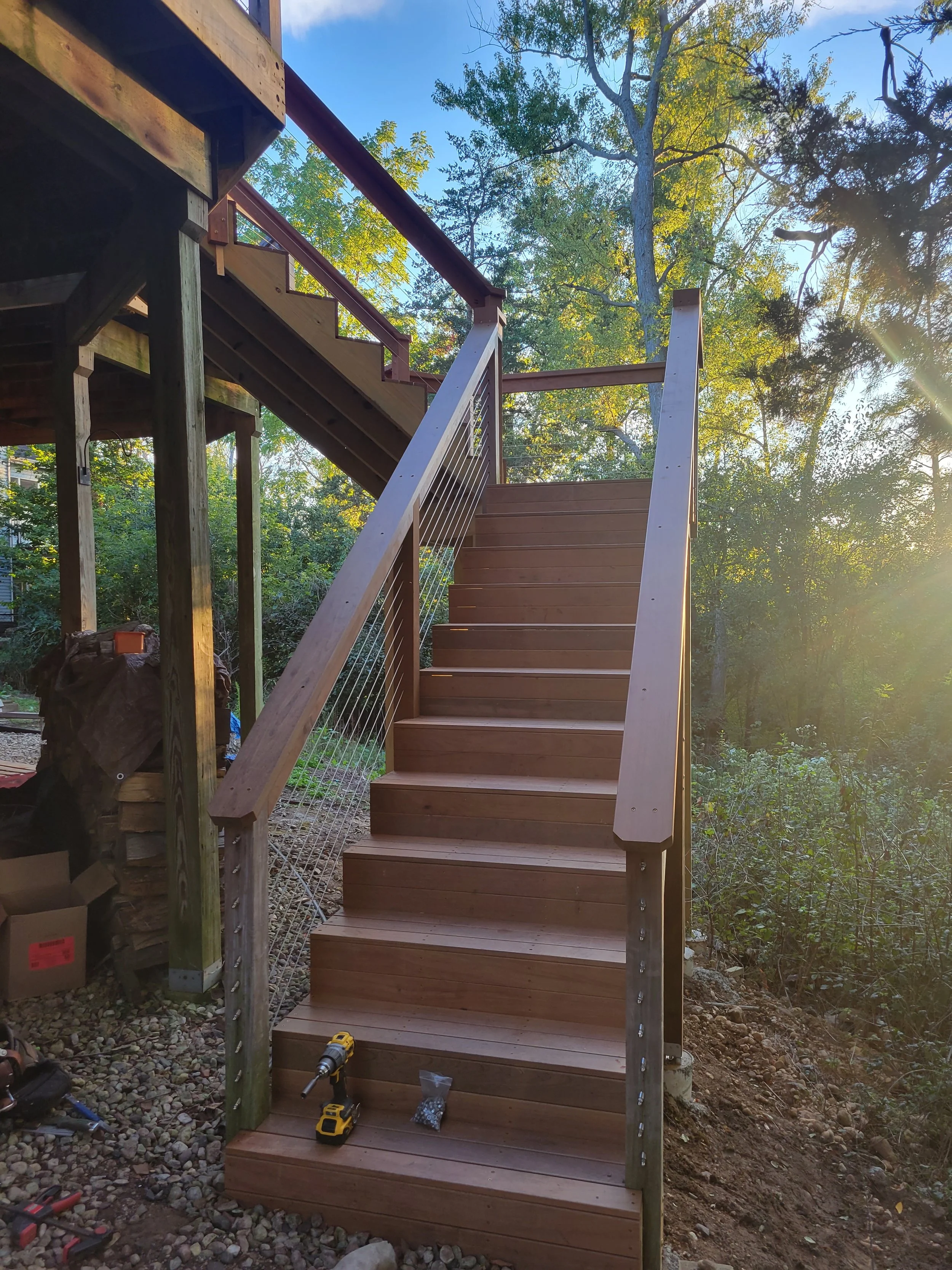 Wooden outdoor staircase with metal railing under construction, located in a lush, green, wooded area during sunset.