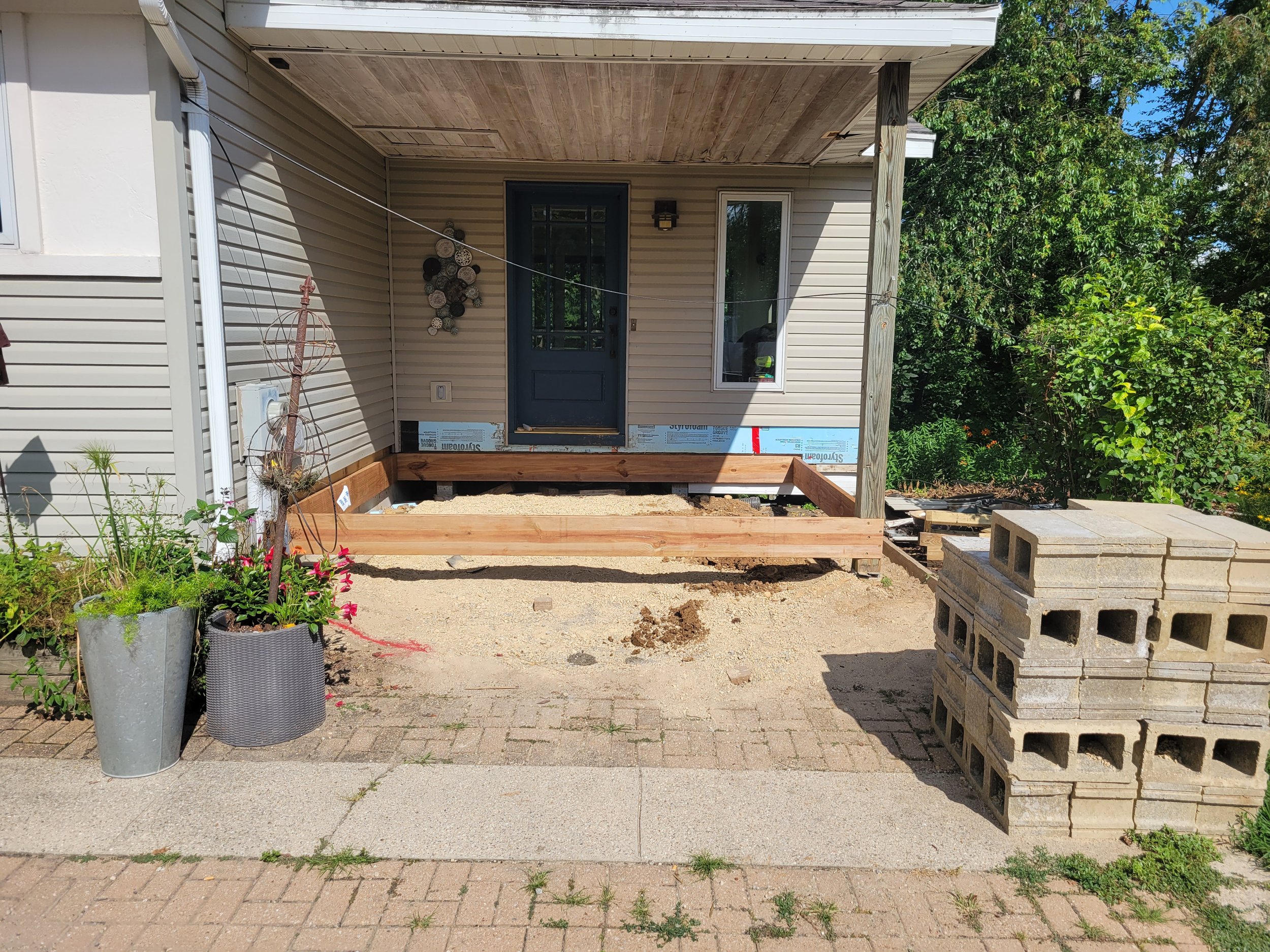 Front porch under construction with wooden framing, construction materials, and potted plants nearby.