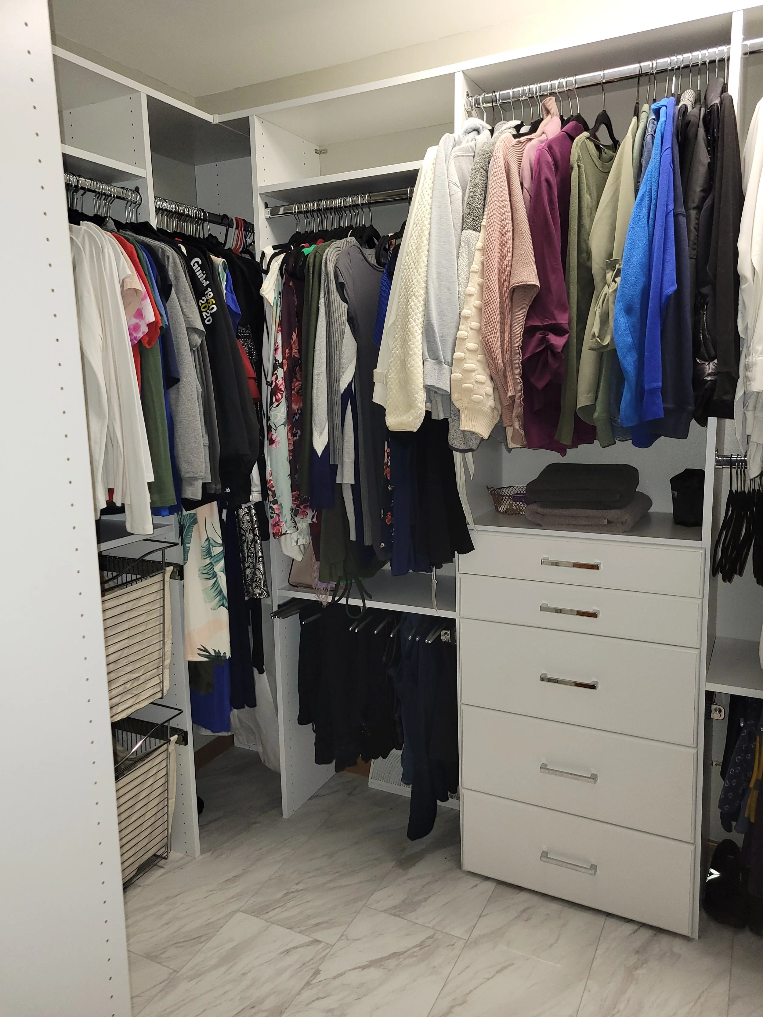 Organized walk-in closet with hanging clothes on rods, shelves, and drawers, featuring shirts, sweaters, and pants, with storage baskets and folded clothes.